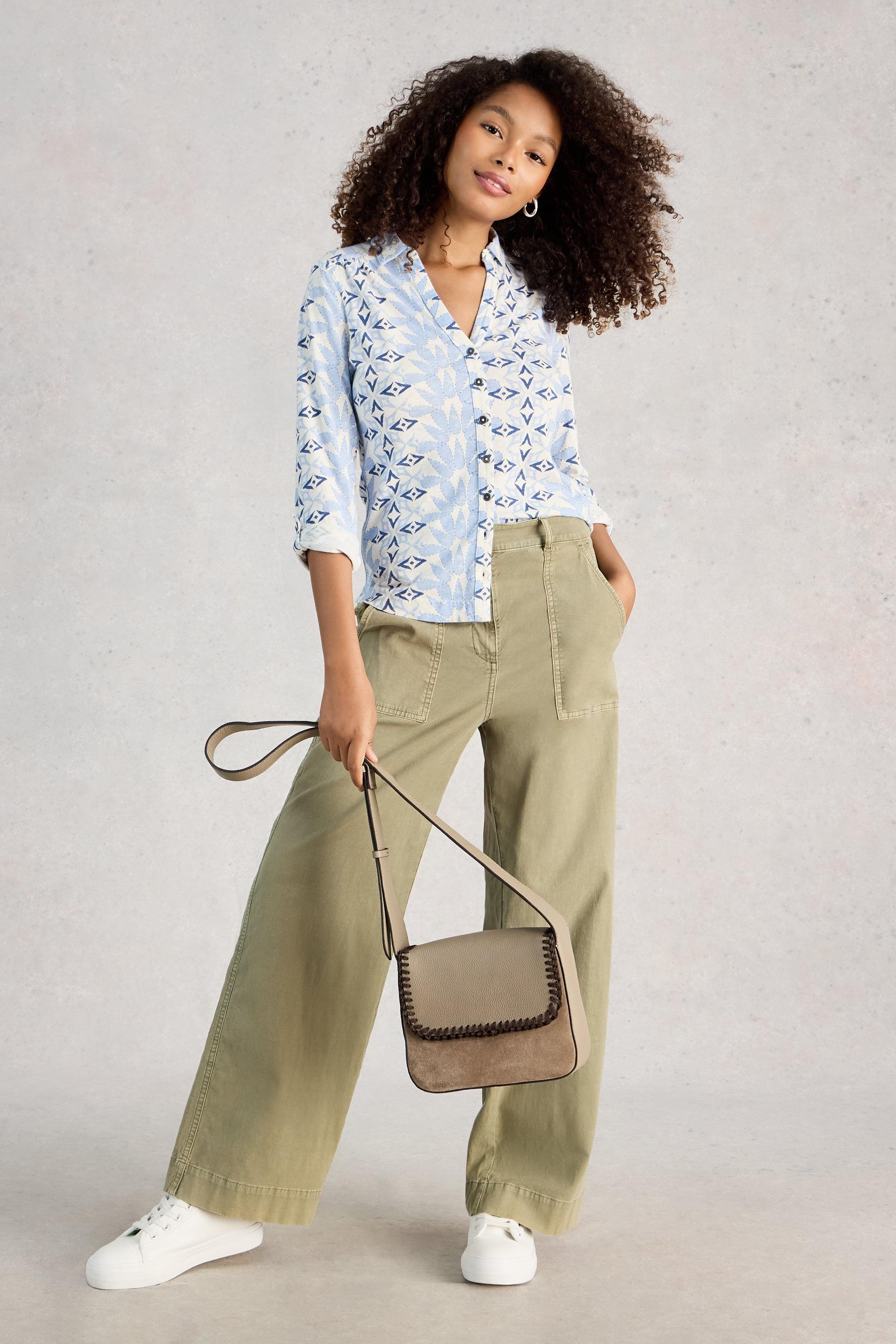 Woman wearing a light blue patterned shirt and green pants, holding a beige handbag against a plain background.