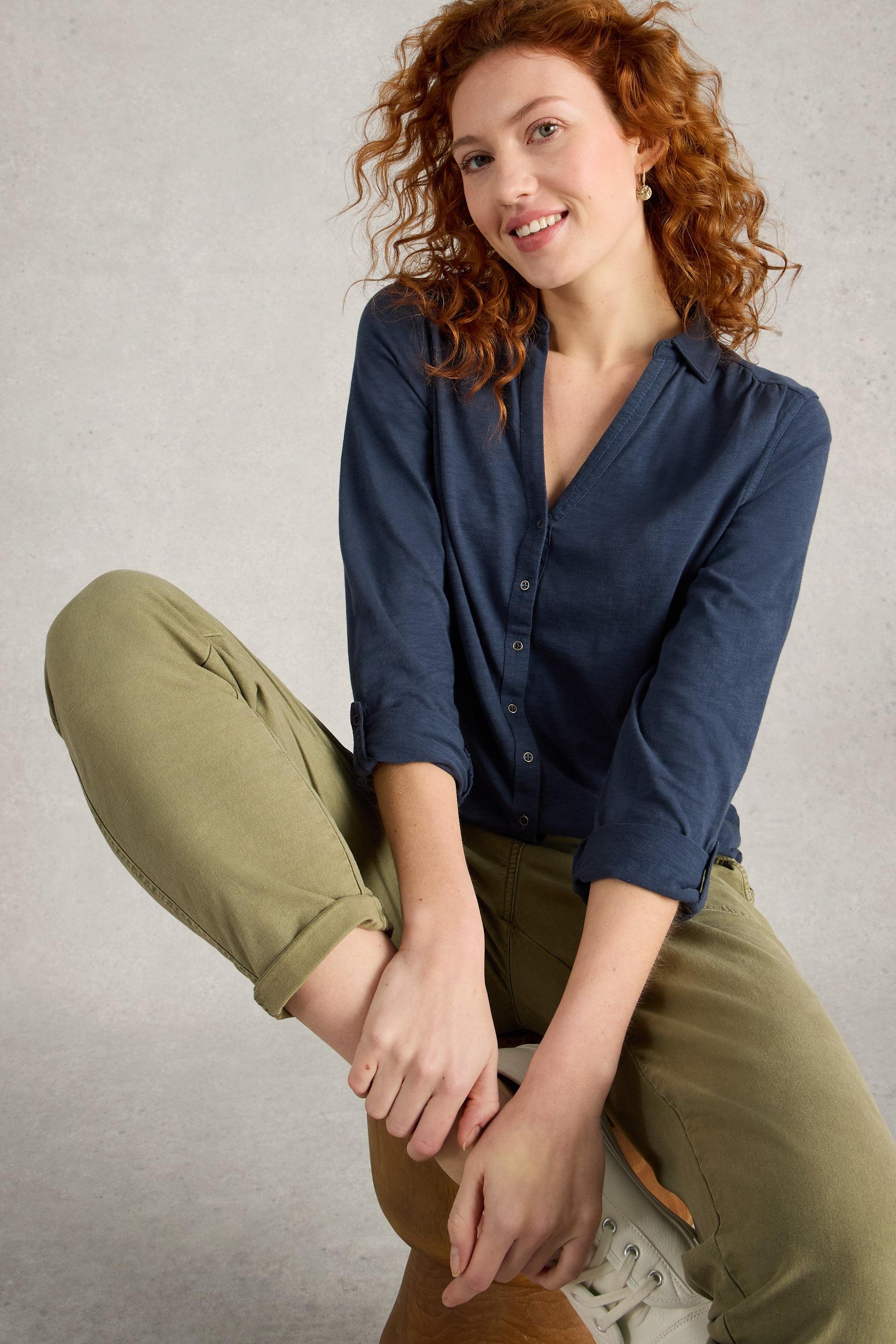 Woman wearing a blue cotton shirt and green pants sitting on a wooden stool against a gray background