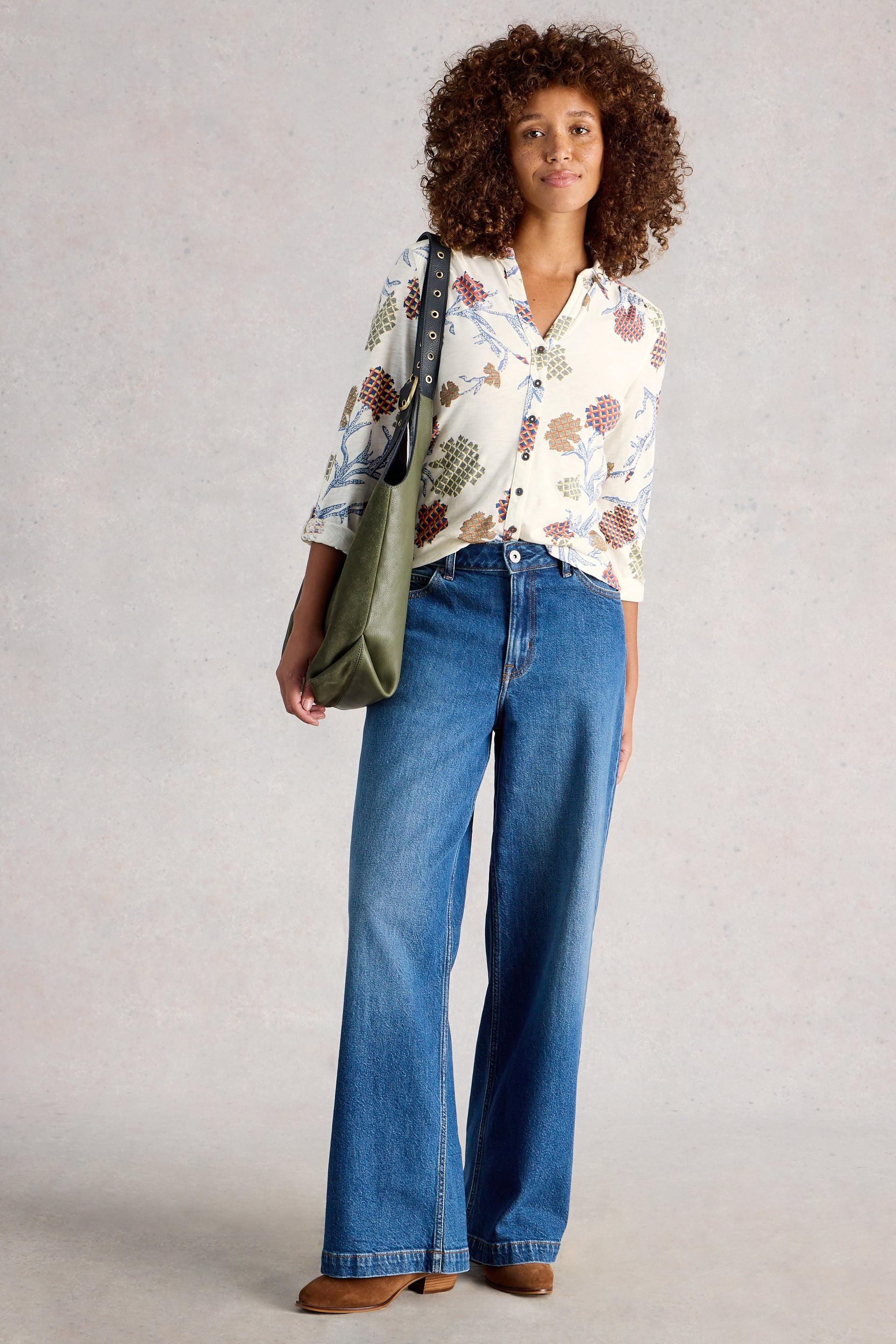 Person wearing a floral cotton blouse and blue jeans, holding a green bag on a plain background