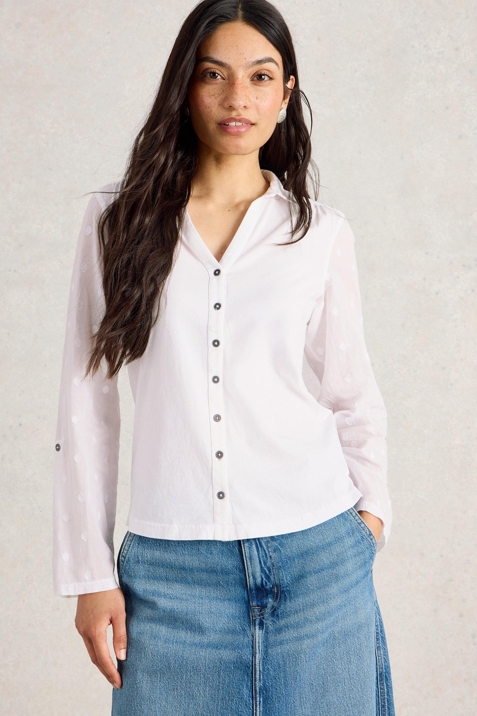 Woman wearing a white cotton blouse with black buttons and blue jeans against a plain background