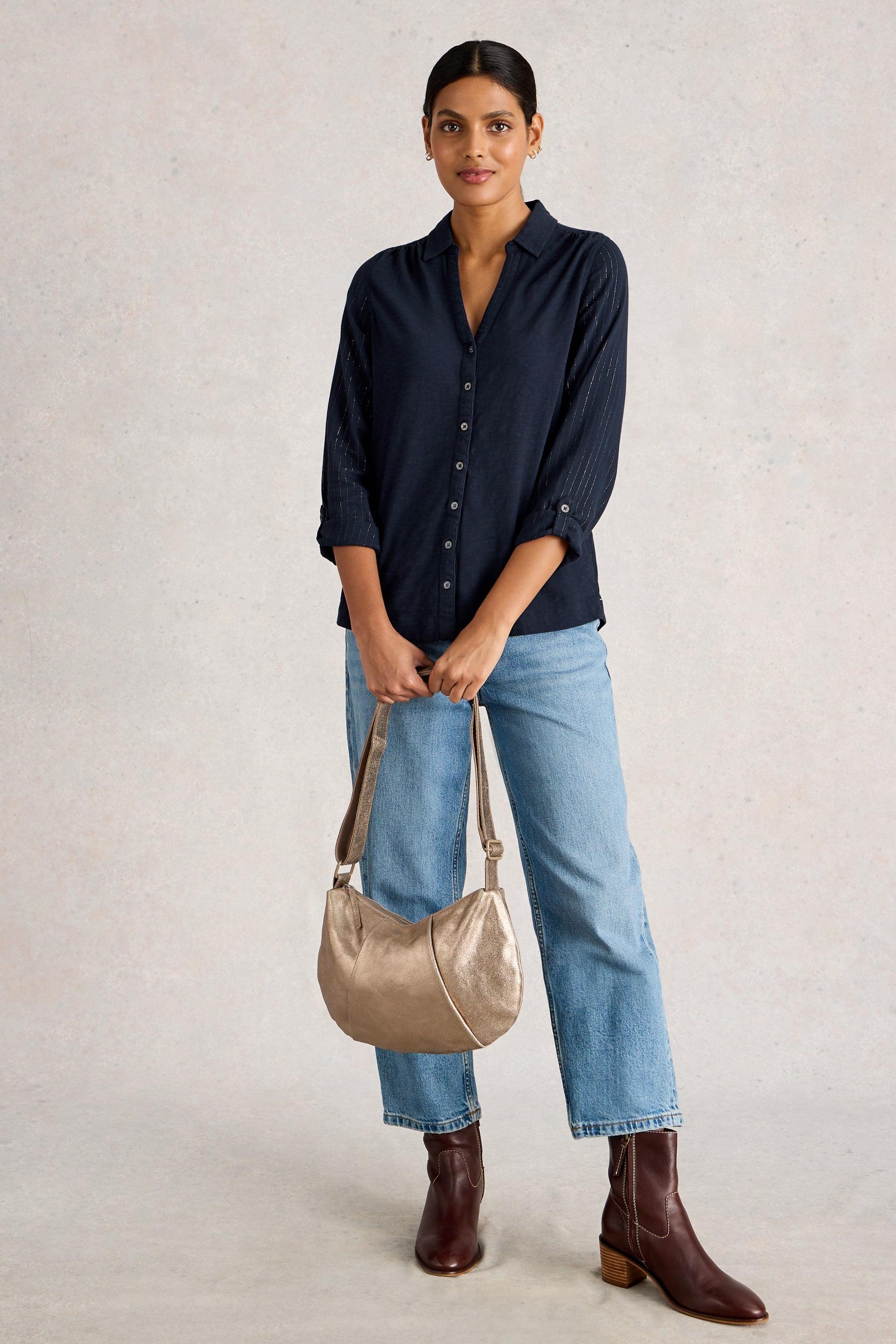 Woman wearing a navy shirt, light blue jeans, and brown boots, holding a beige handbag against a plain background.