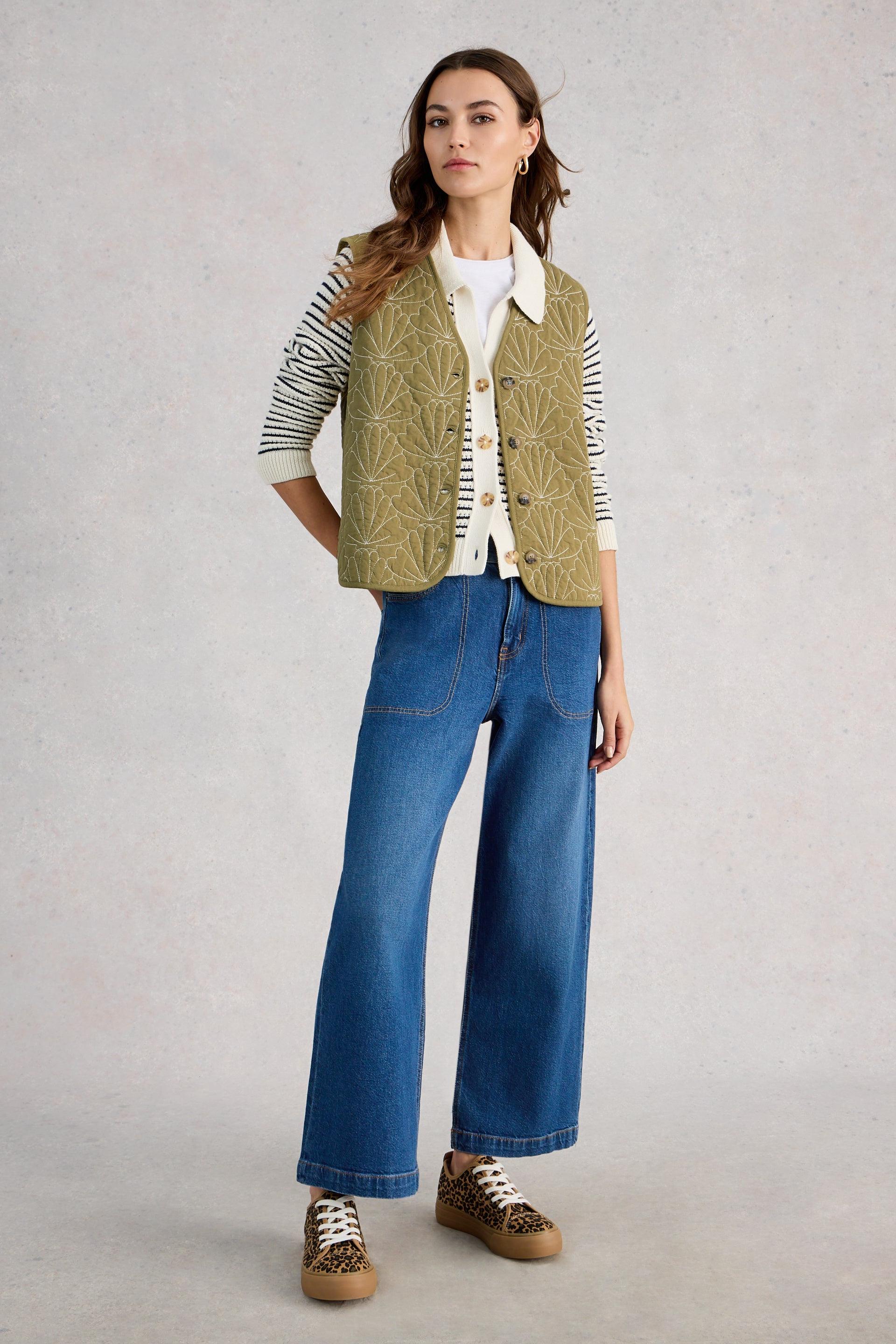 Woman wearing a green embroidered gilet, white shirt, blue jeans, and brown shoes on a light gray background