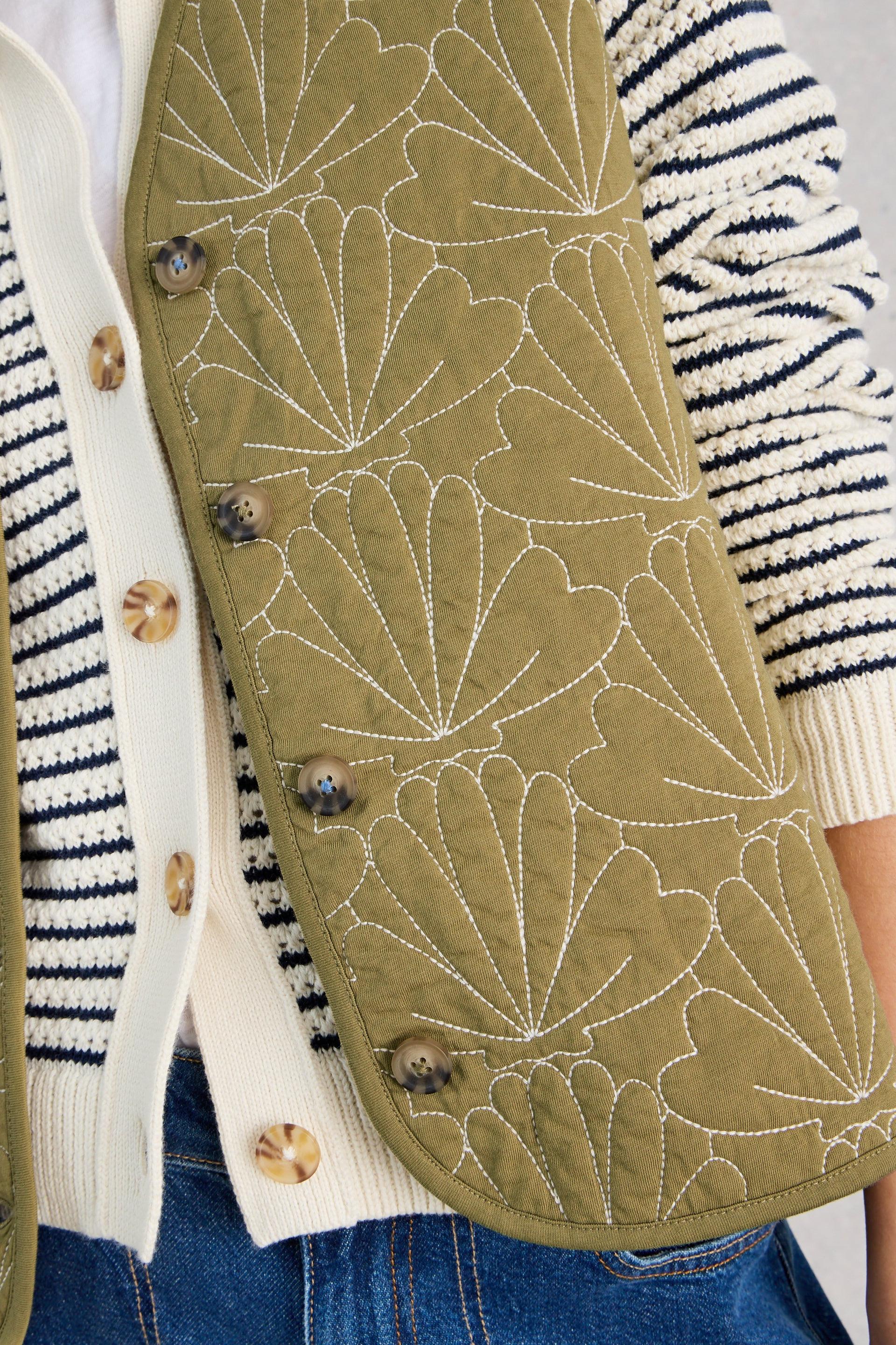 Close-up of a green quilted vest with white floral patterns worn over a striped sweater and jeans.
