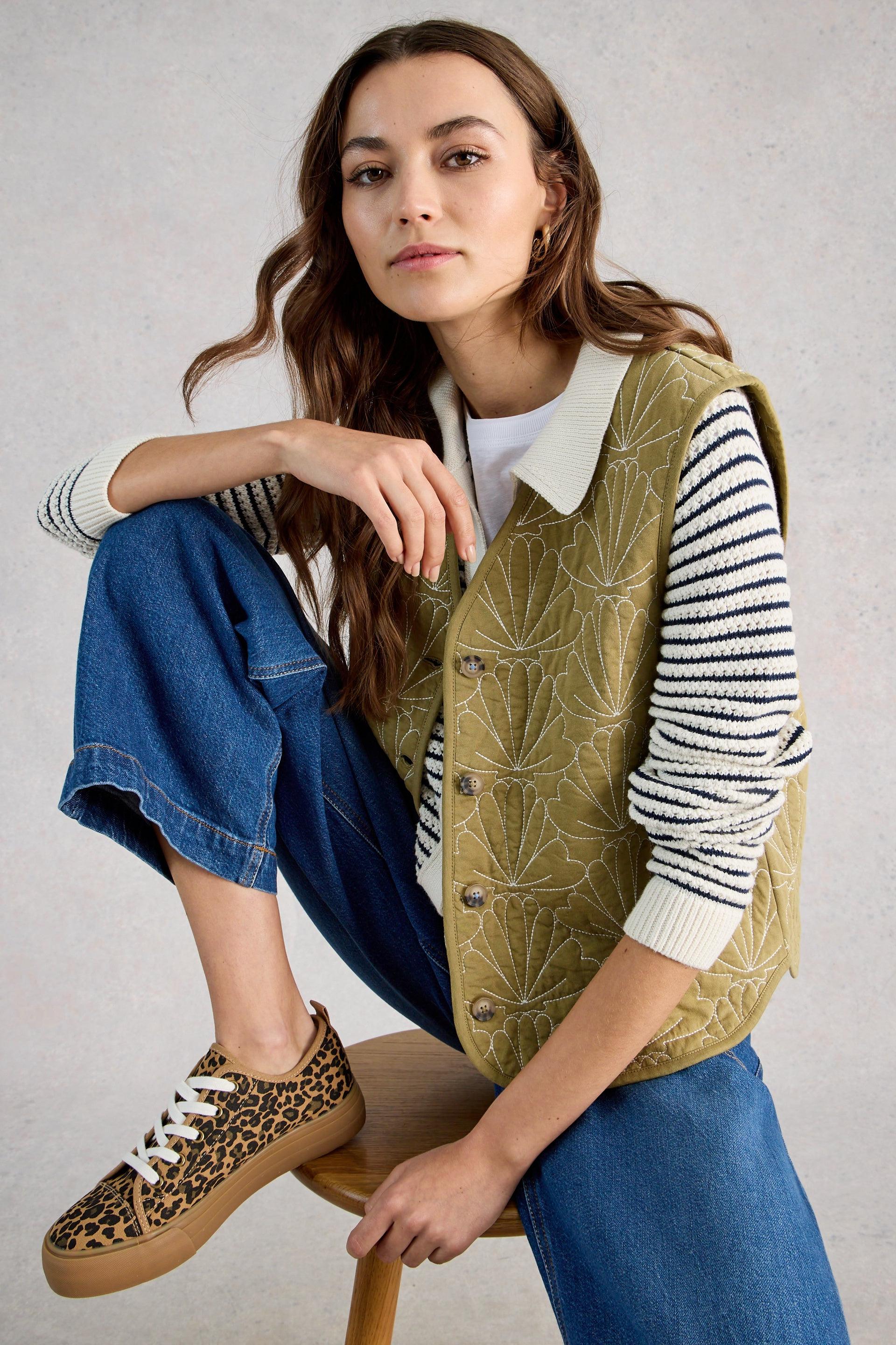 Woman wearing a green quilted gilet striped sweater, and leopard print shoes sitting on a wooden stool.