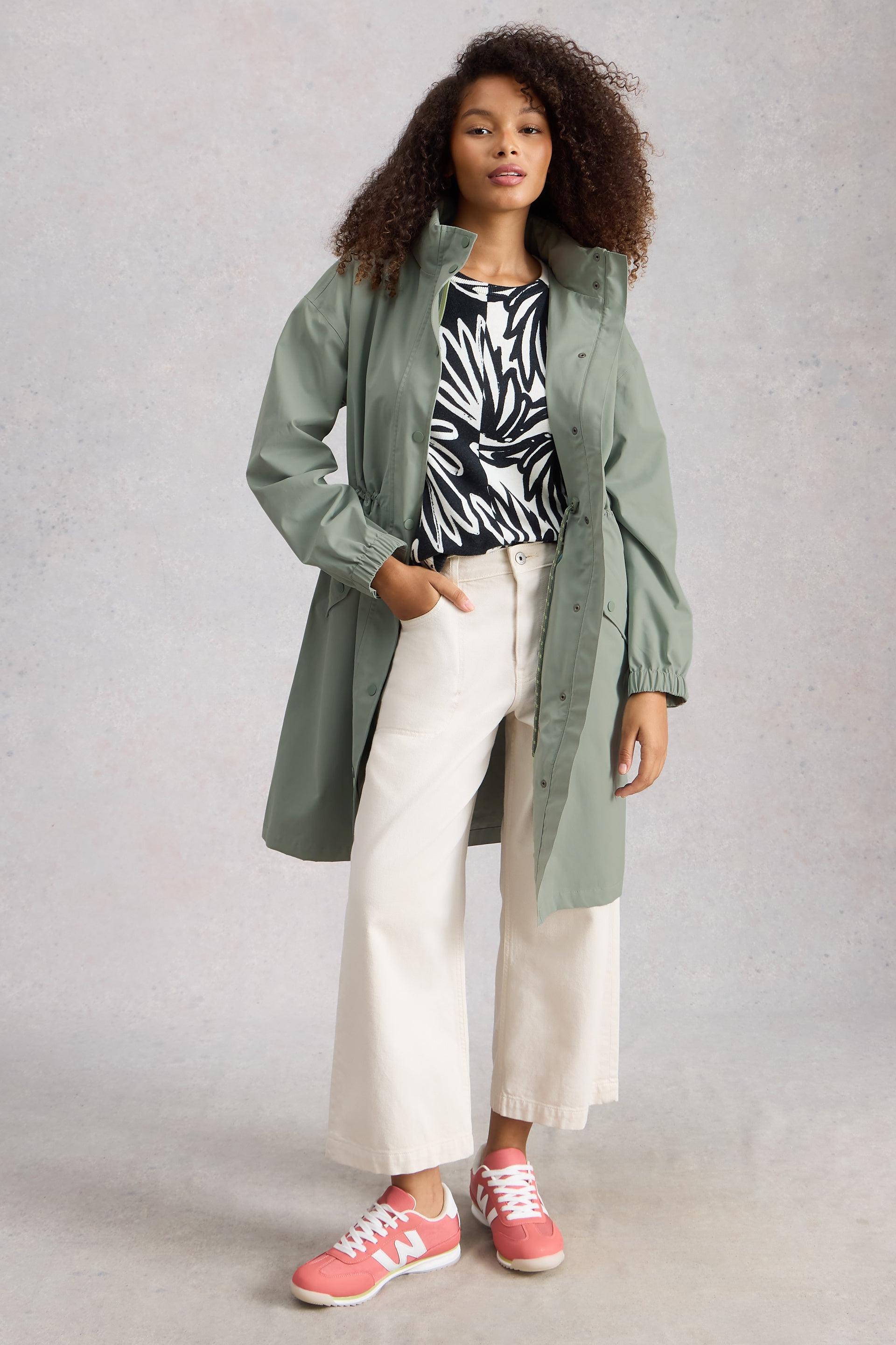 Woman wearing a green waterproof coat, zebra print top, white pants, and pink sneakers against a plain background