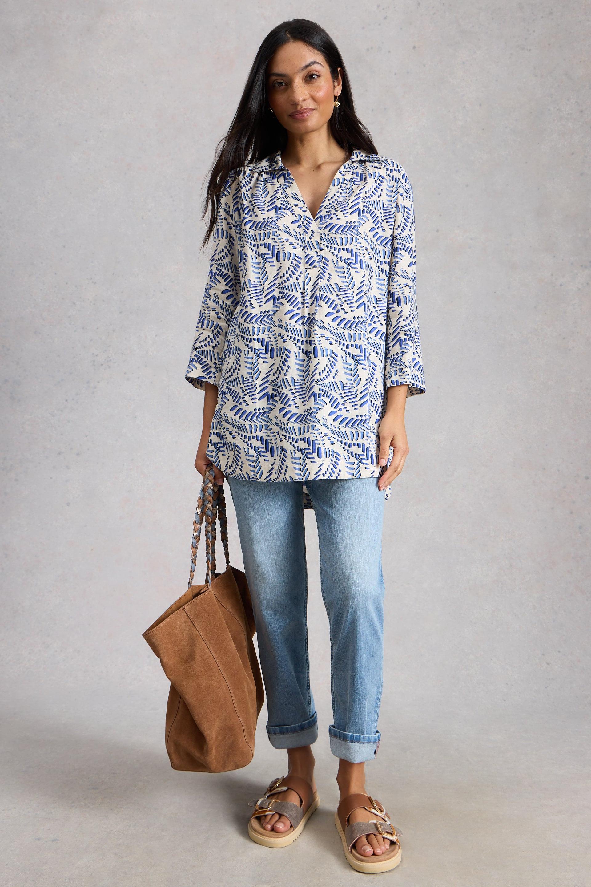 Woman wearing a blue organic cotton tunic and jeans, holding a brown bag on a gray background