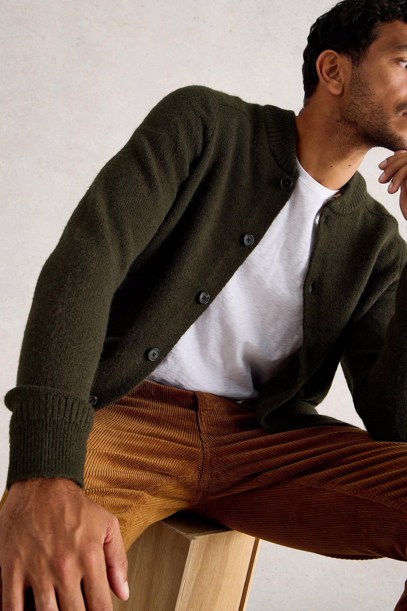 Man sitting on wooden box wearing woolen bomber cardigan with white t-shirt and orange ribbed trousers