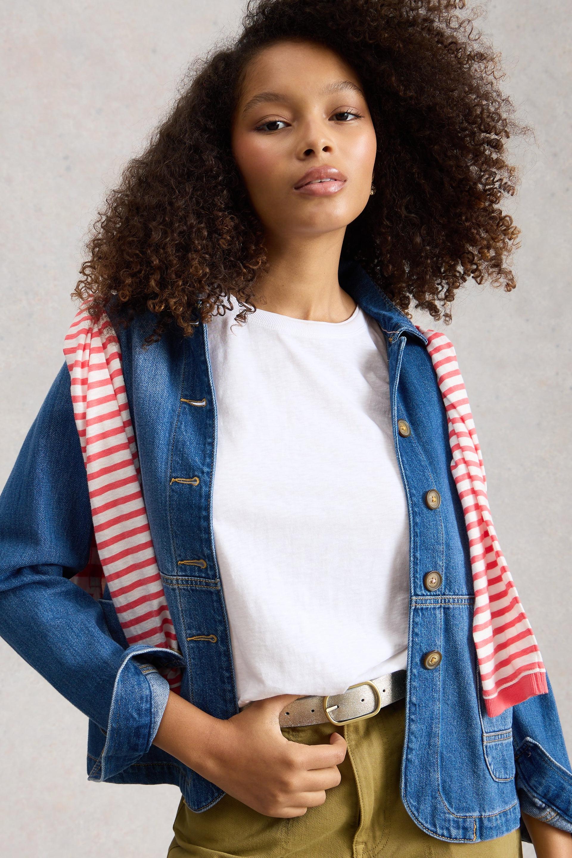 Person wearing a denim jacket over a white t-shirt with a red and white striped shirt around her shoulders, on a neutral background.
