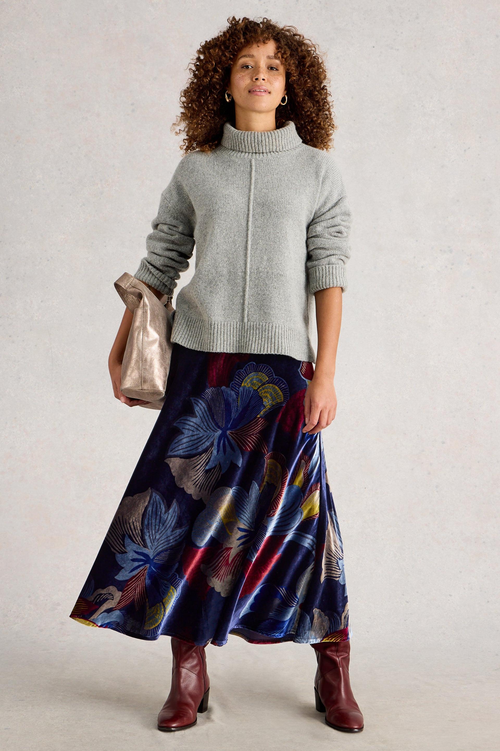 Woman wearing a gray sweater and colorful floral skirt with red boots, holding a silver handbag.