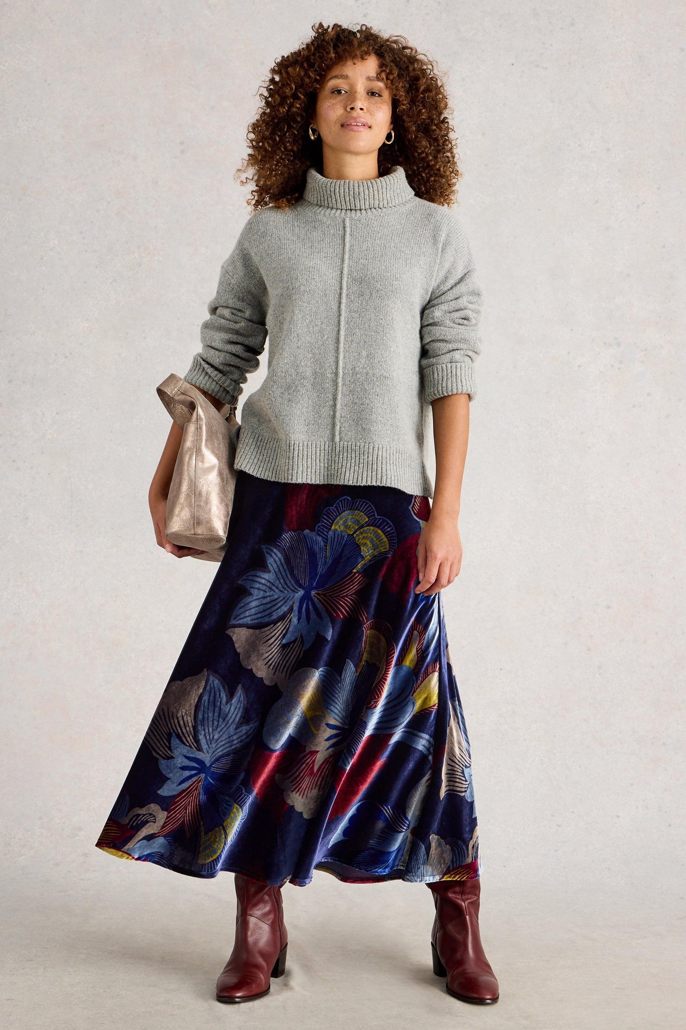 Woman wearing a gray sweater and colorful floral skirt with red boots, holding a silver handbag.