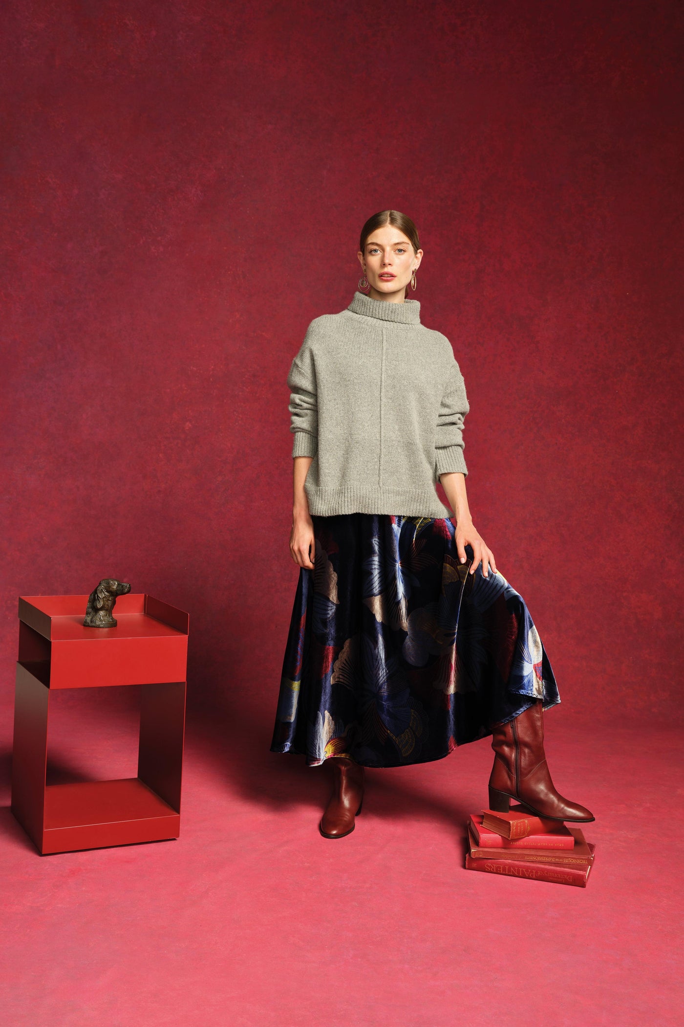 Woman in a gray sweater and dark skirt standing next to a red table against a red background