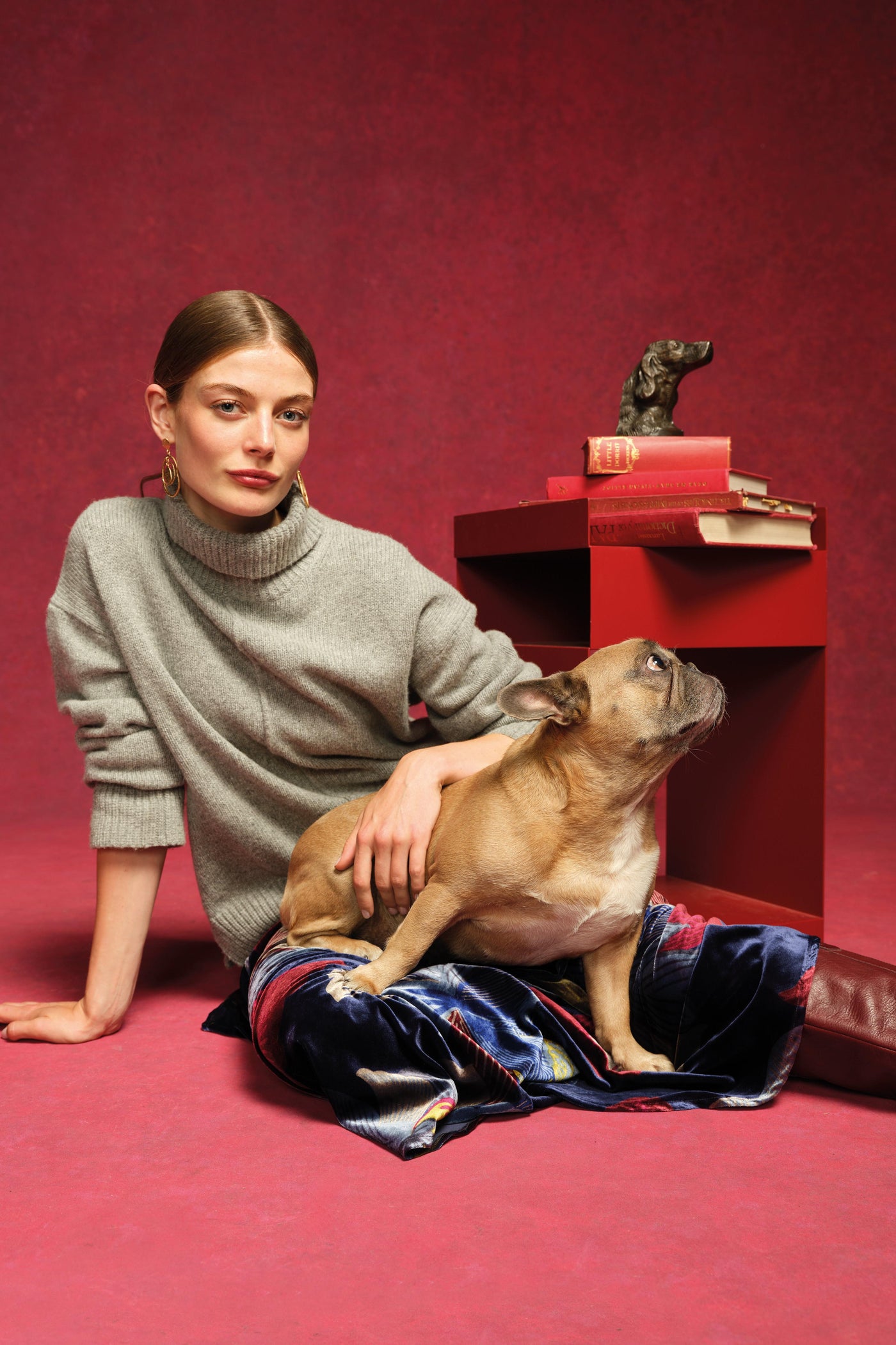 Woman in a gray sweater sitting on a red floor with a dog, against a red wall.