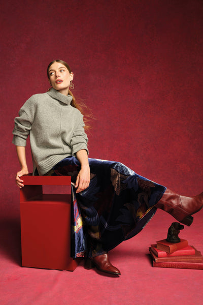 Woman in a gray sweater and blue skirt sitting on a red stool against a red background