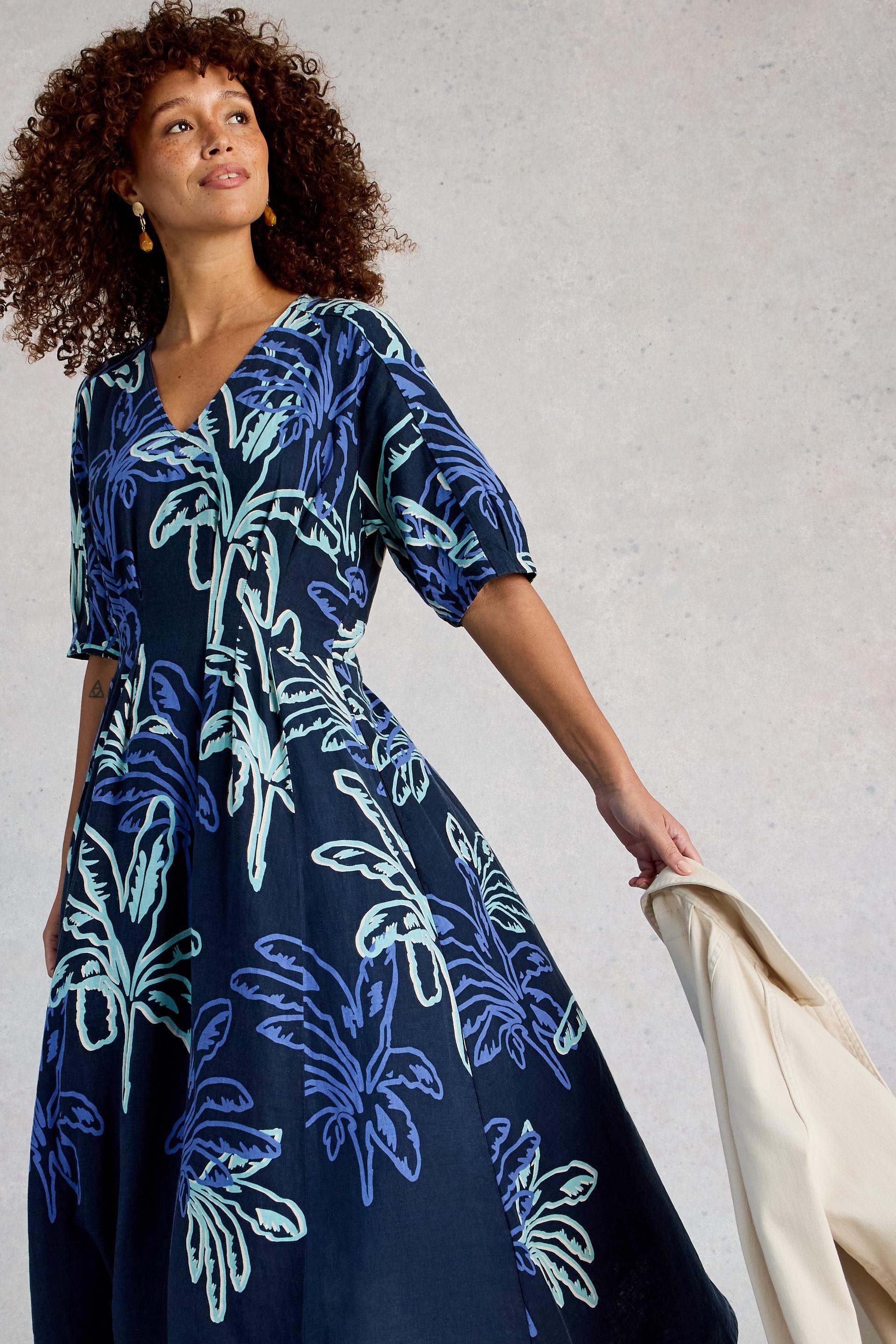 Woman wearing a navy linen dress with blue floral patterns, holding a beige jacket against a neutral background.
