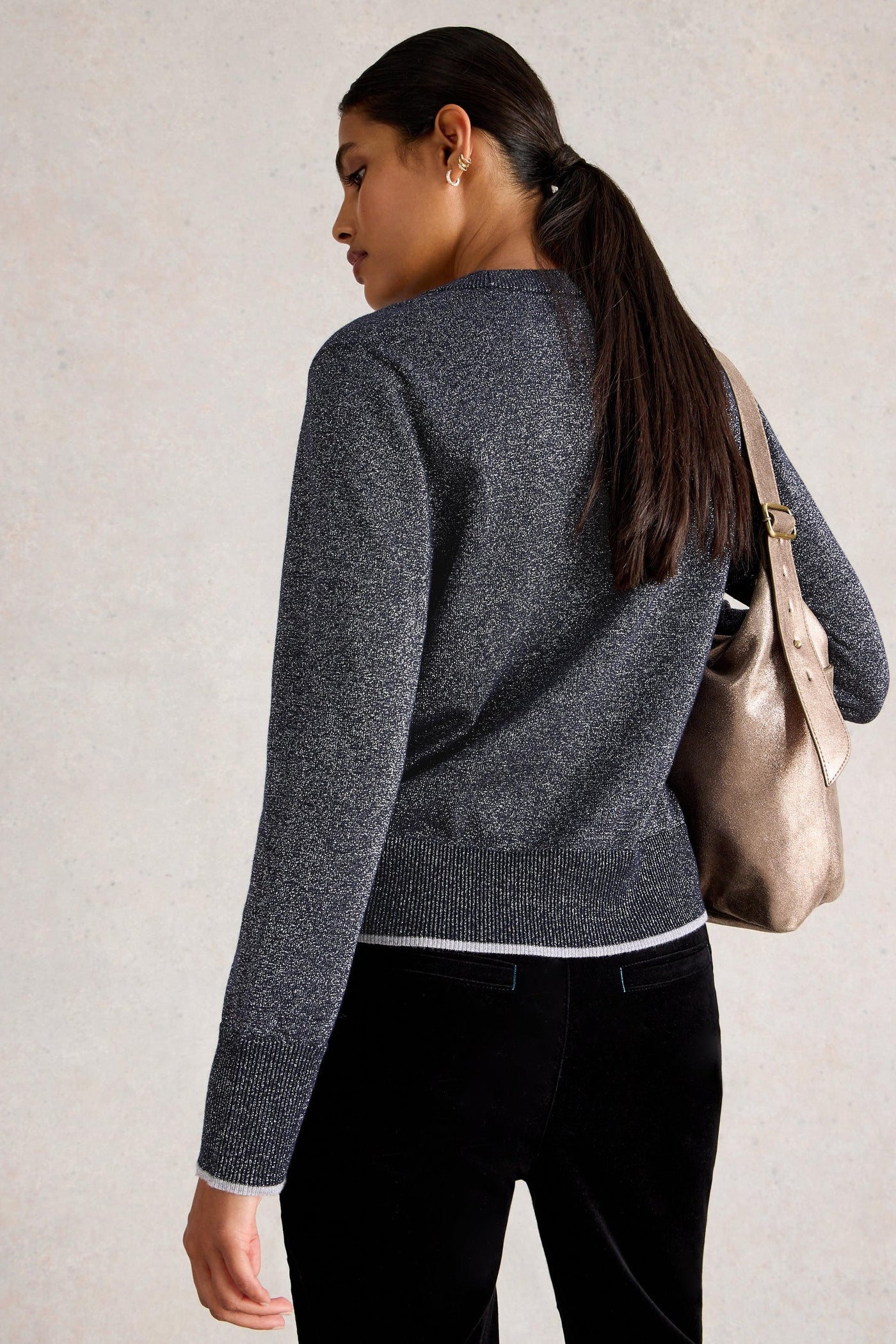 Woman wearing a gray organic cotton sweater and black pants, carrying a brown metallic bag on a plain background