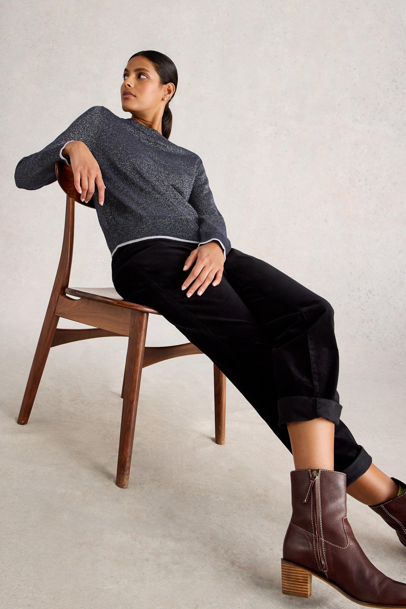 Woman sitting on a wooden chair wearing a gray organic cotton sweater, black pants, and brown boots.