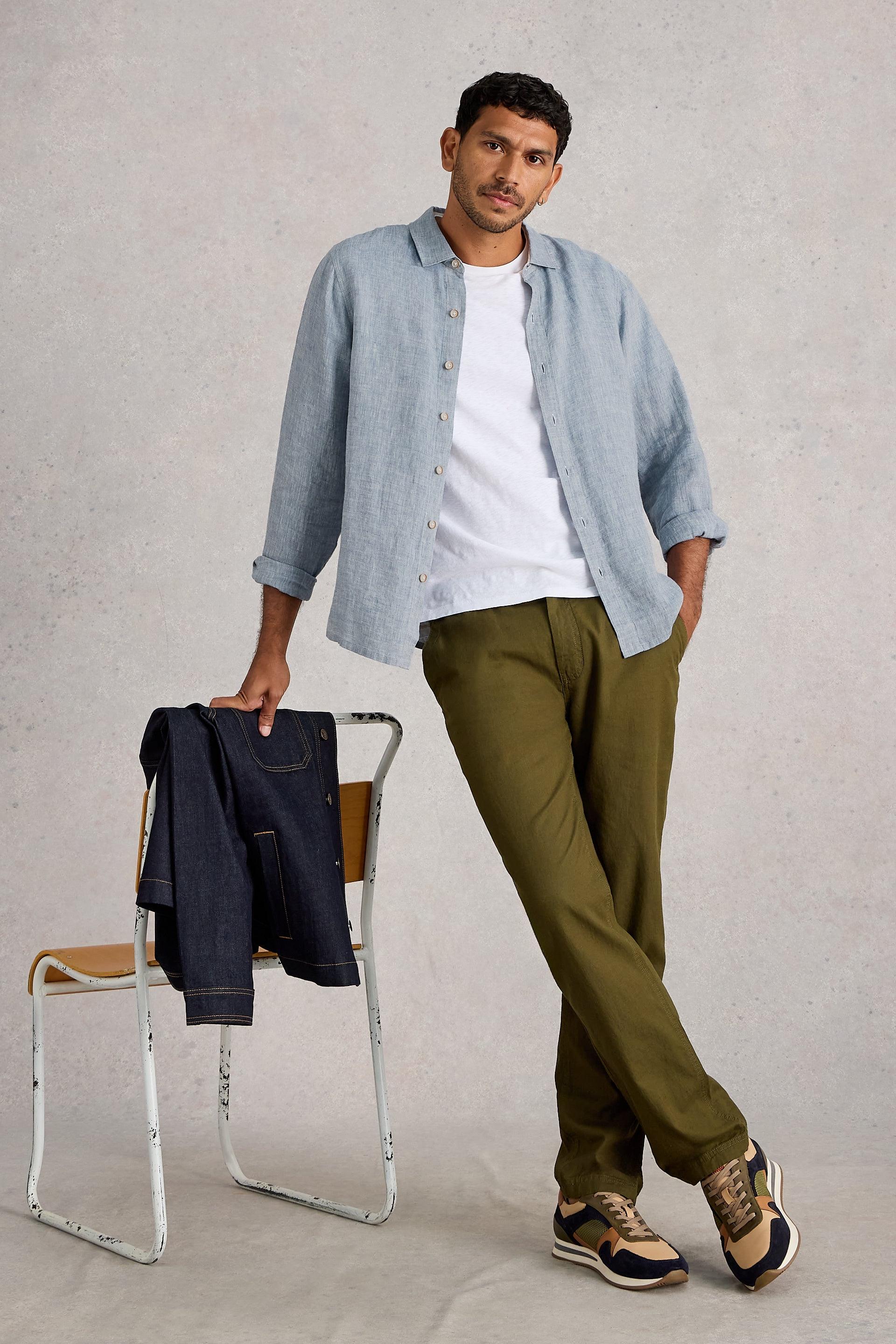 Man wearing a light blue linen shirt, white t-shirt, and green pants, standing next to a chair with folded jeans on it.