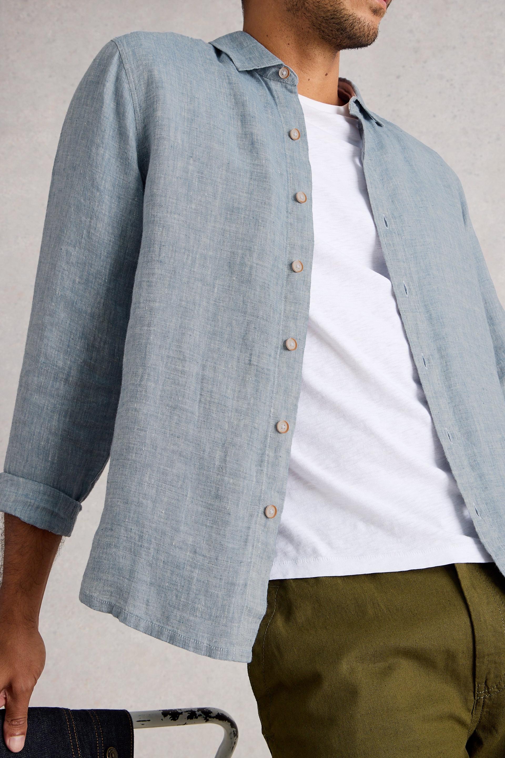 Person wearing a light blue button-up shirt over a white t-shirt with green pants on a plain background