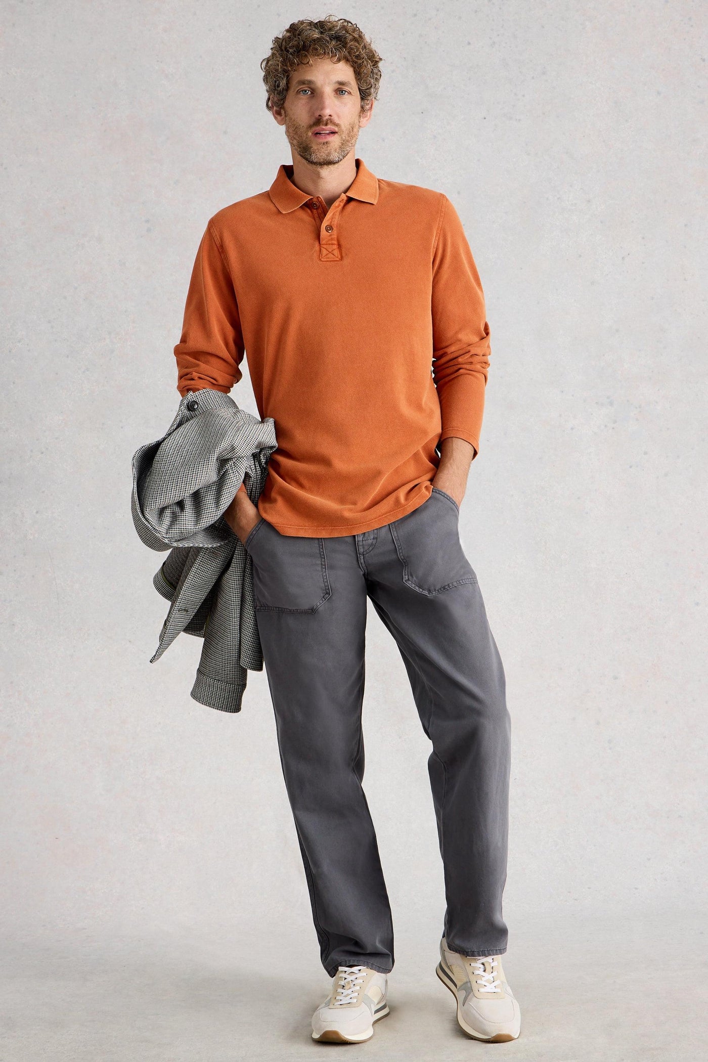 Man standing wearing fairtrade cotton long sleeve orange polo styled with grey chino trousers and white sneakers
