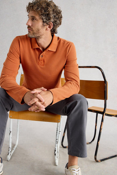 Man sitting on chair wearing fairtrade cotton long sleeve orange polo styled with grey trousers