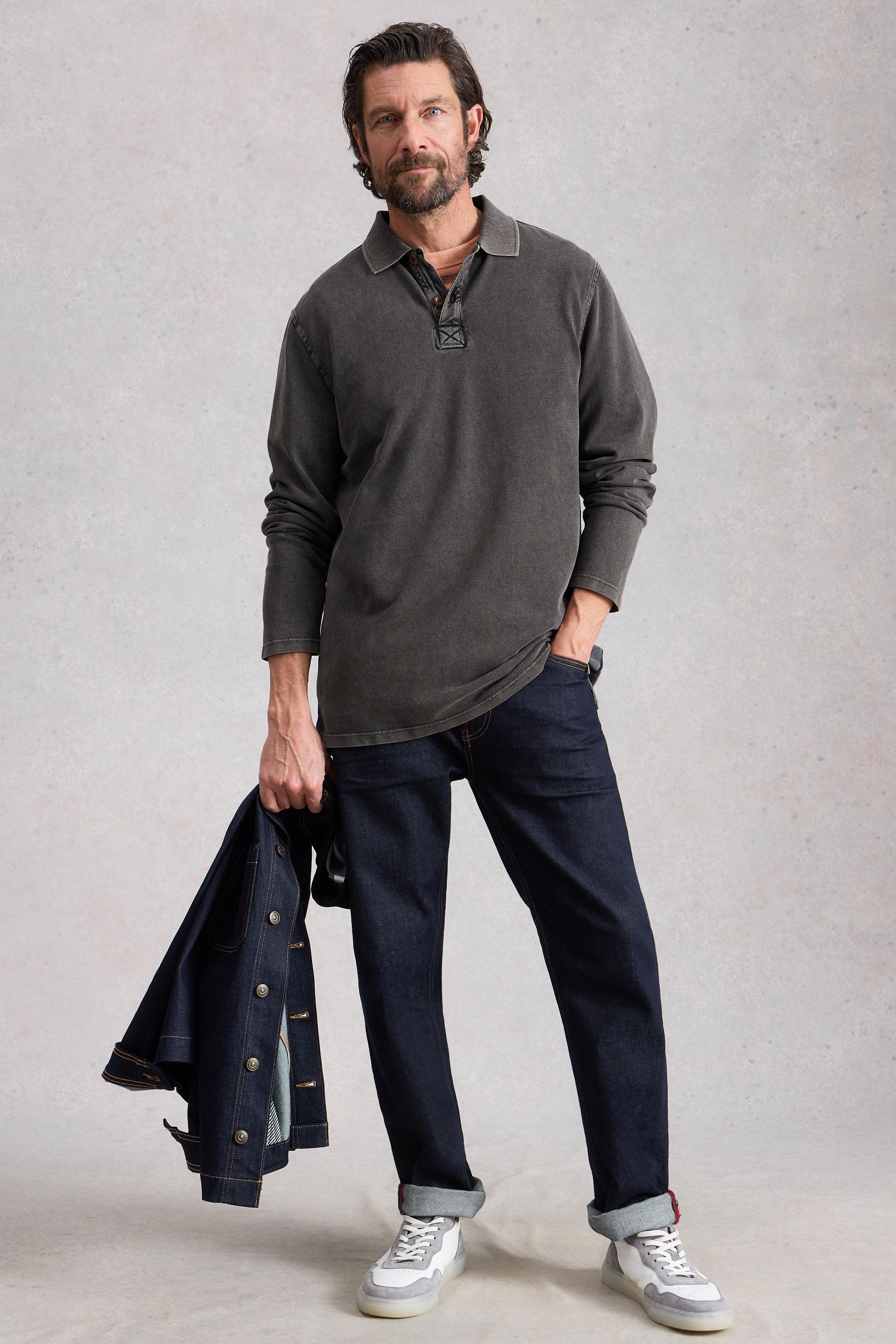 Man wearing a gray long-sleeve shirt and dark jeans, holding a denim jacket, against a plain background.