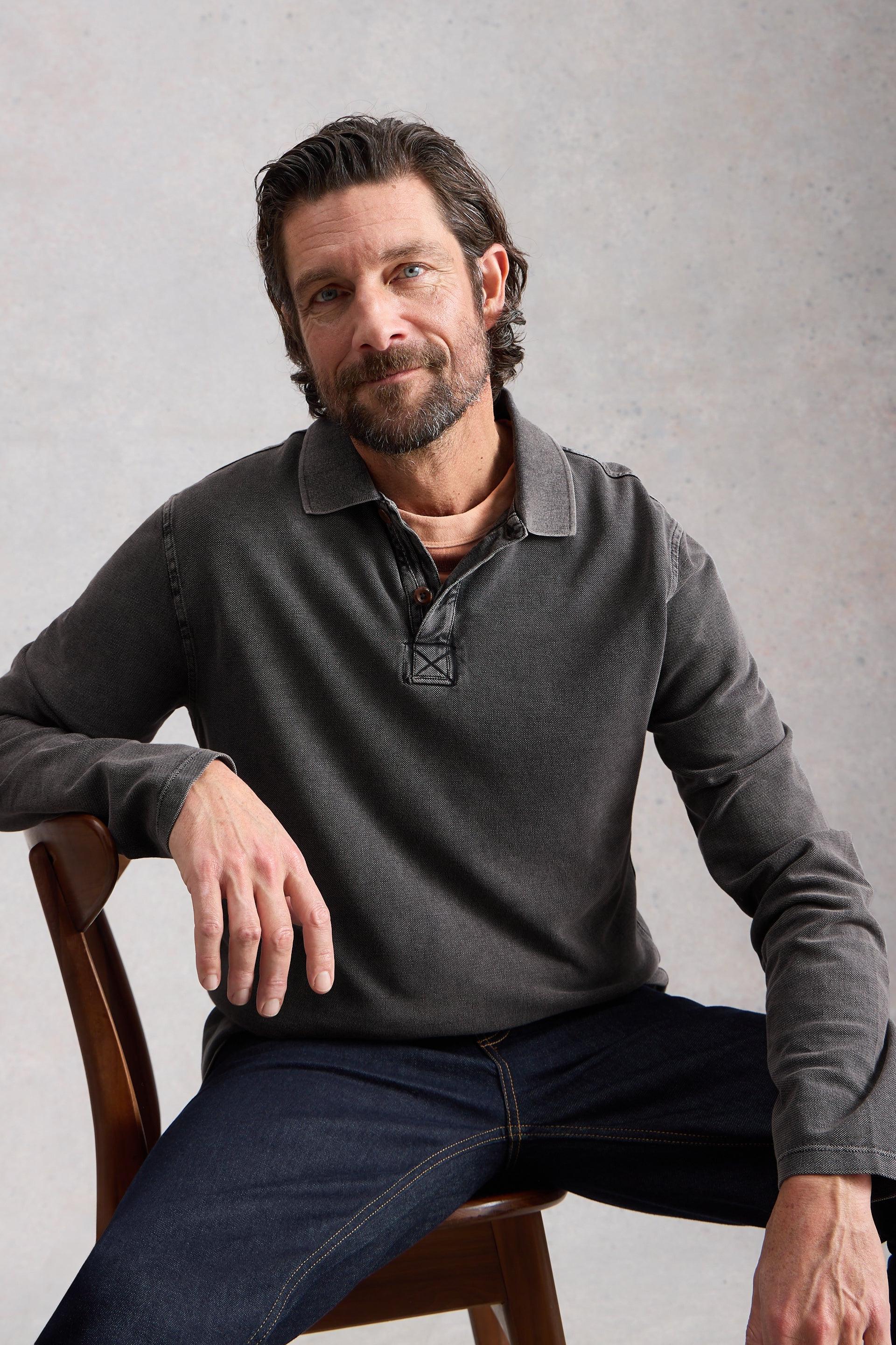 Man sitting on a wooden chair wearing a gray cotton sweater and dark jeans against a plain background