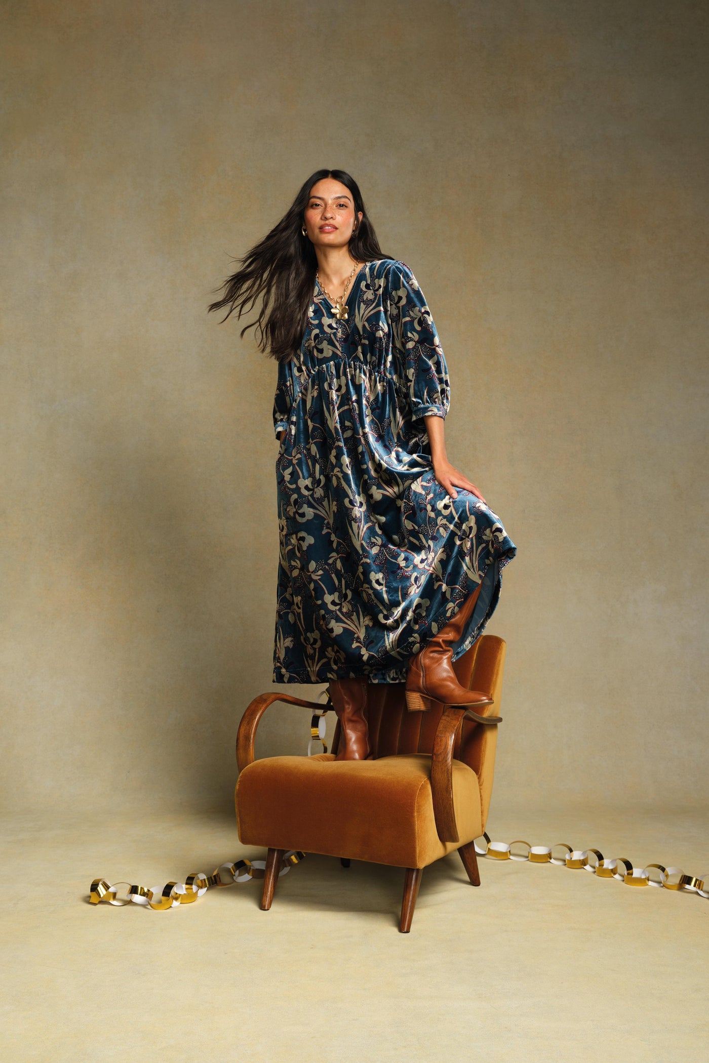 Woman in a blue patterned dress standing next to an orange chair against a beige background