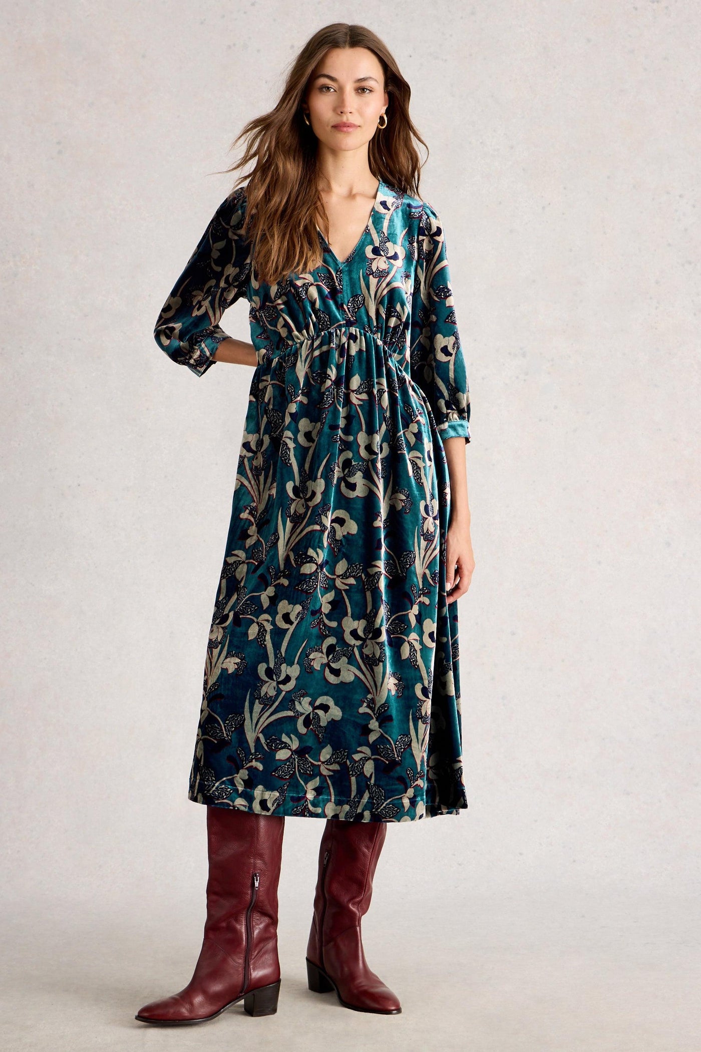 Woman wearing a floral velvet dress with long sleeves and burgundy boots on a plain background