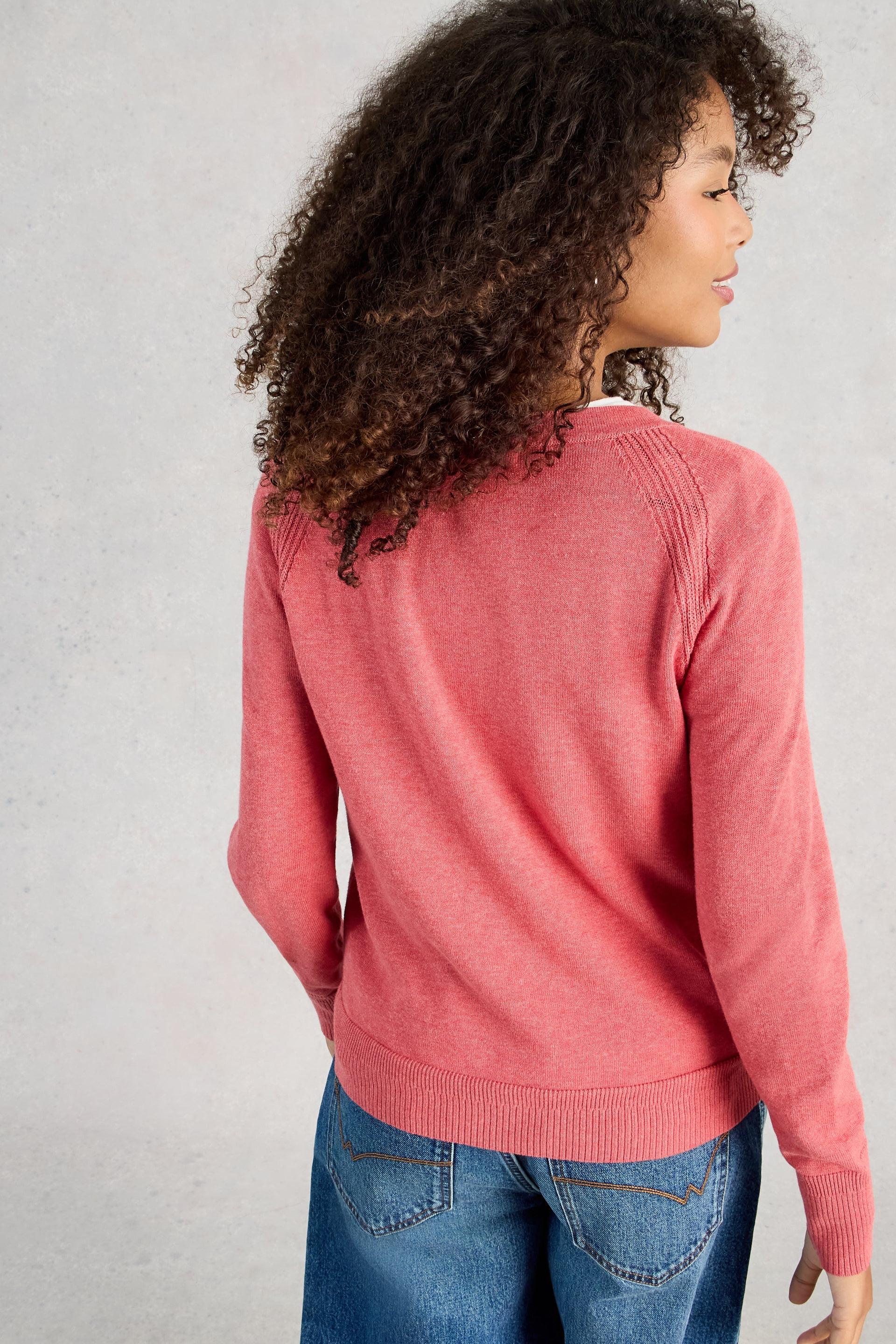 Woman wearing a pink organic cotton cardigan and blue jeans against a plain background