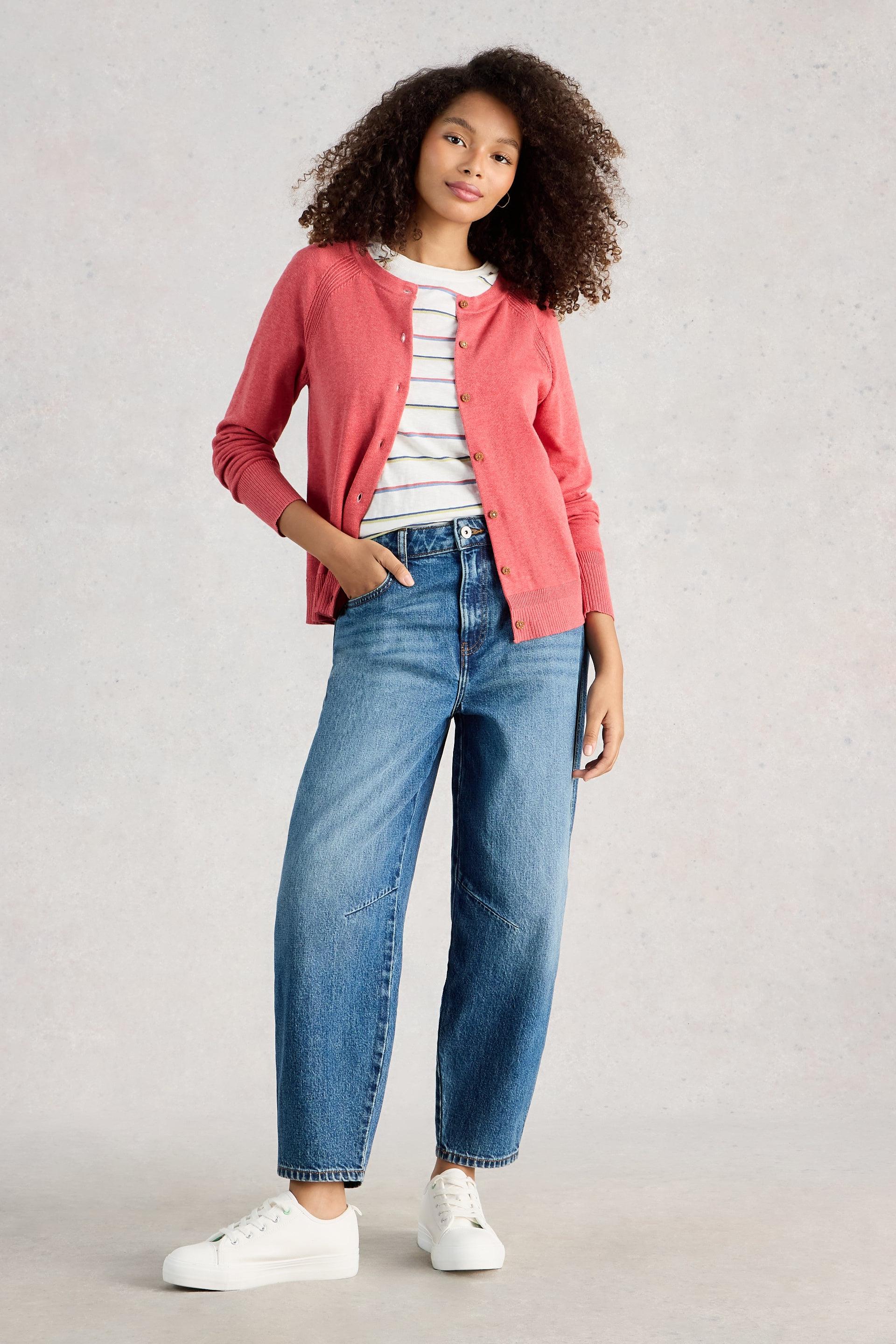Model wearing a pink organic cotton cardigan, striped shirt, blue jeans, and white sneakers on a light gray background
