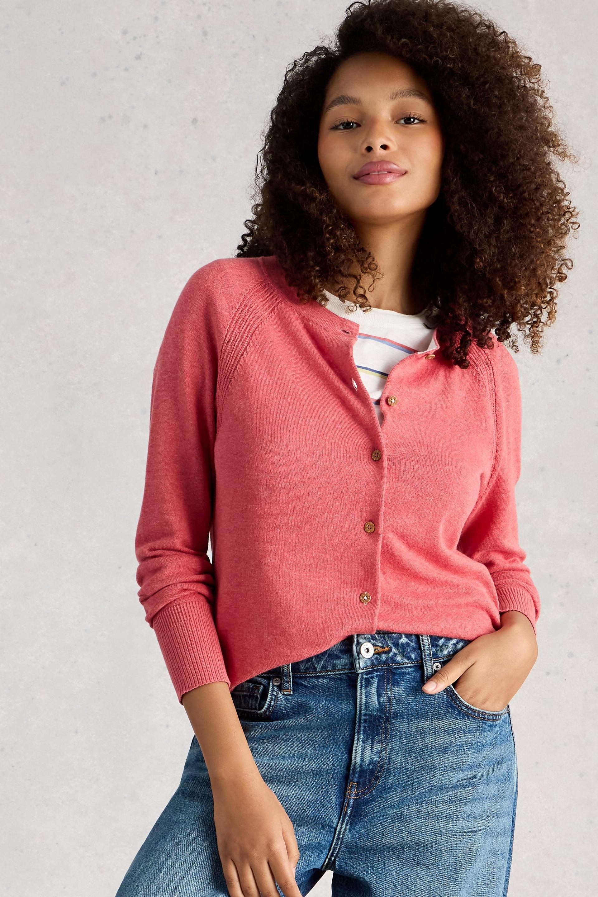 Woman wearing a pink organic cotton cardigan over a striped shirt with blue jeans against a light gray background