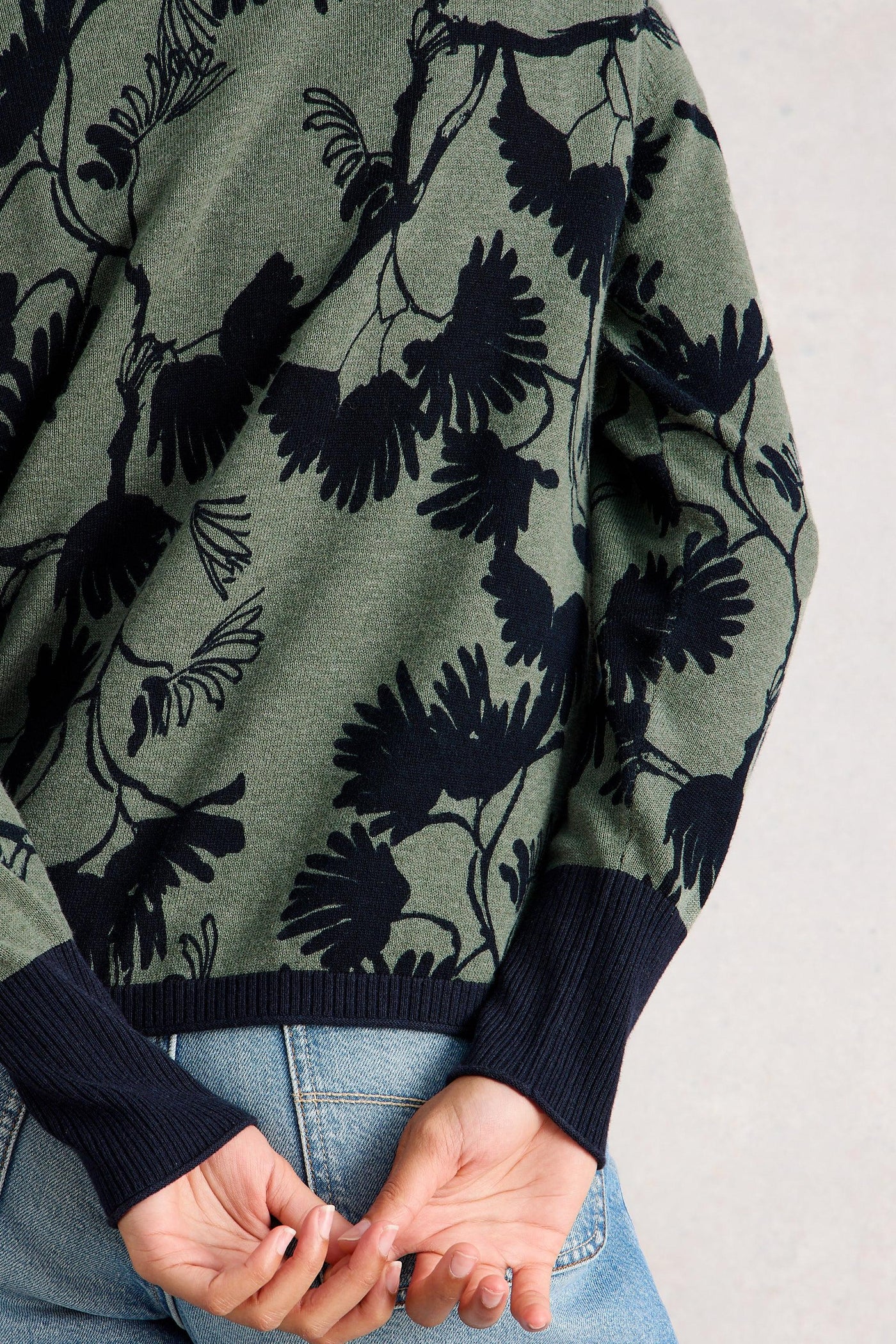 Back view of a green and navy floral organic cotton cardigan with ribbed navy cuffs and hem, worn with light blue jeans, hands clasped behind the back.