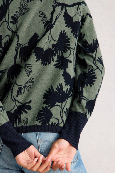Back view of a green and navy floral organic cotton cardigan with ribbed navy cuffs and hem, worn with light blue jeans, hands clasped behind the back.