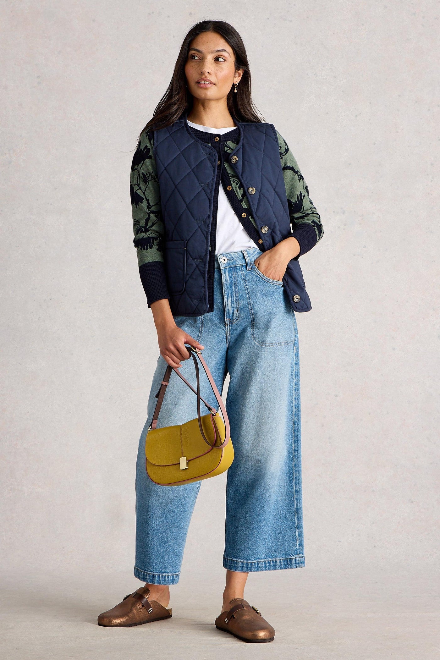 Woman standing wearing a green and navy floral organic cotton cardigan layered under a navy quilted vest, paired with light blue wide-leg jeans and brown loafers, holding a mustard handbag.