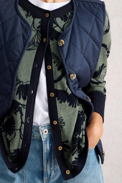 Woman wearing a green and navy floral cardigan layered under a navy quilted vest, showing the front button detail and pockets, paired with light blue jeans.