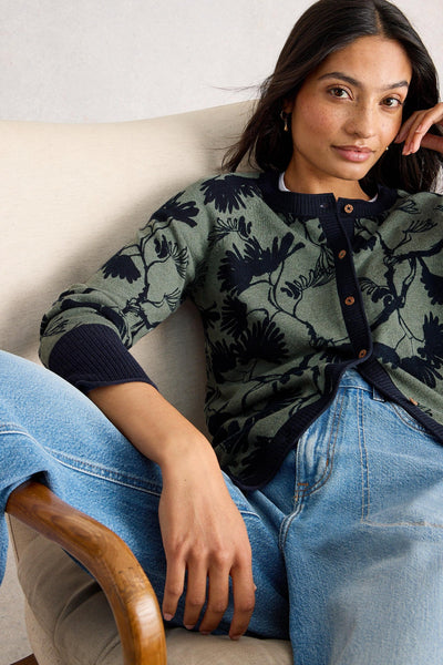 Woman sitting in a beige armchair wearing a green and navy floral organic cotton cardigan with buttons, styled with light blue jeans
