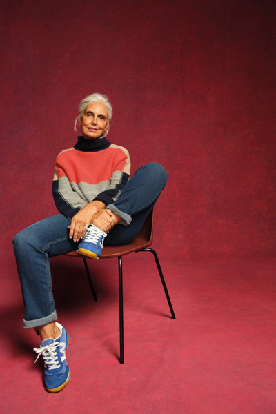 Person sitting on a chair wearing a colourful organic cotton sweater and blue sneakers against a red background
