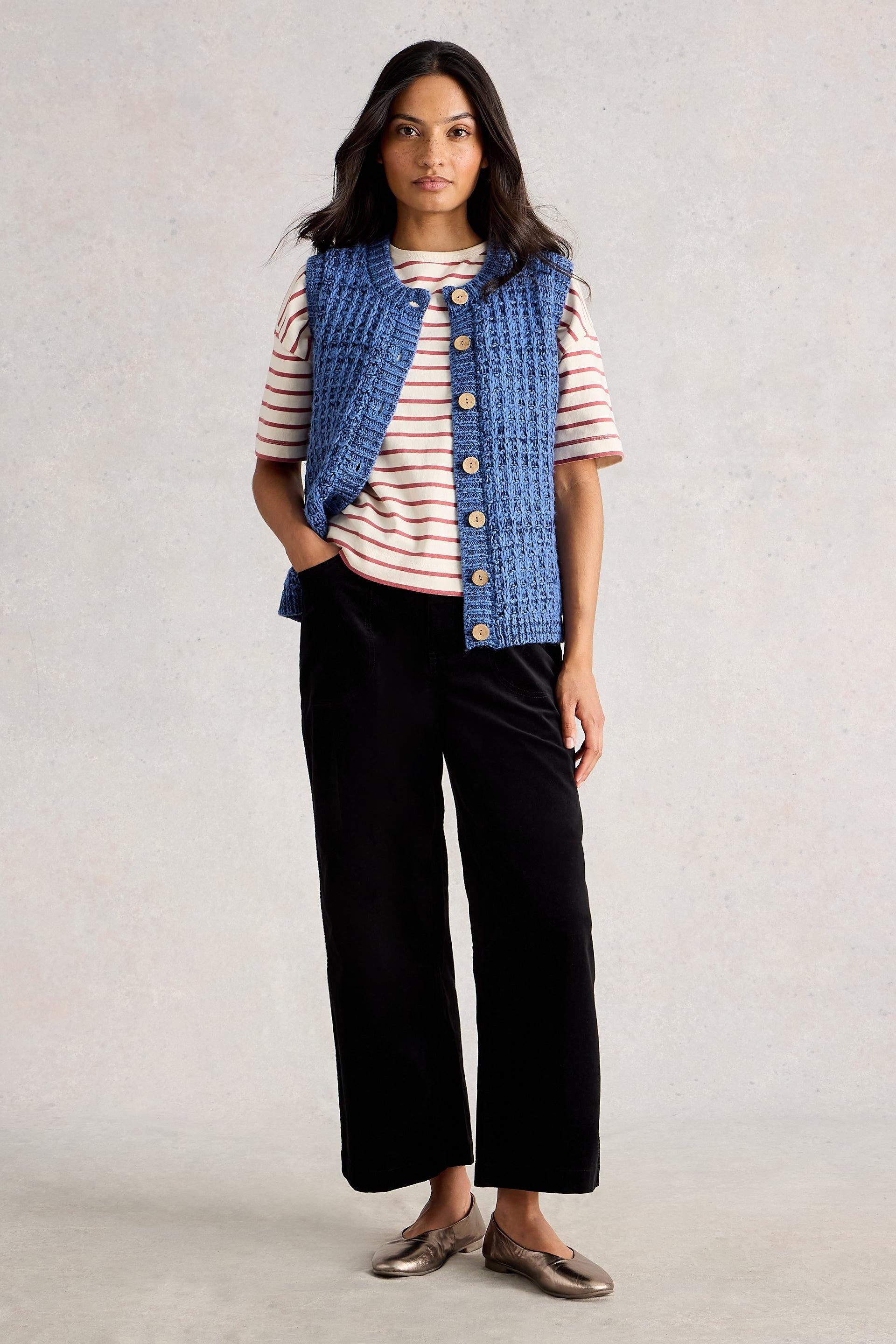 Woman wearing a blue knitted vest over a striped shirt with black pants on a plain background