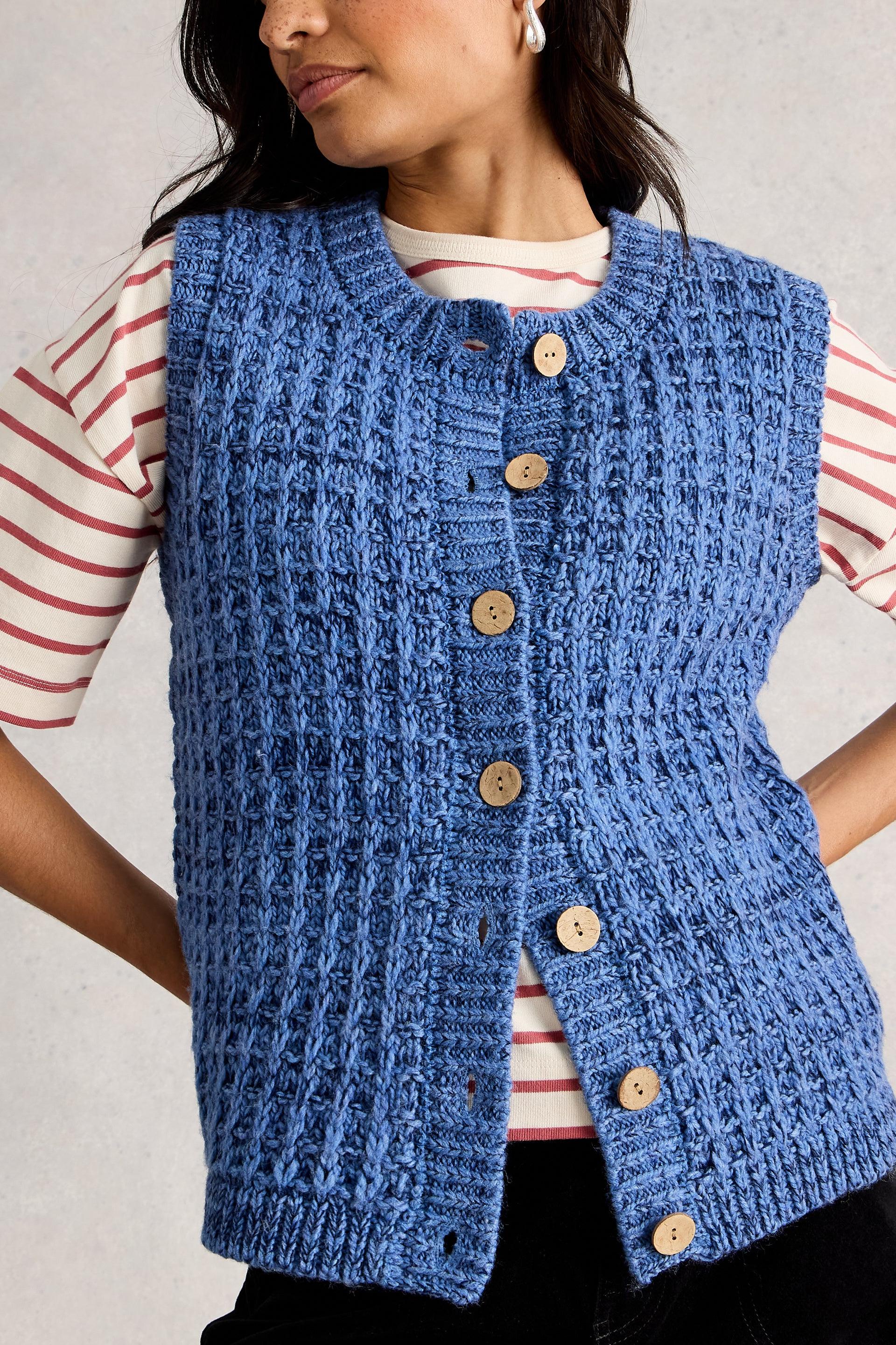 Woman wearing a blue knitted vest with button details on a plain background