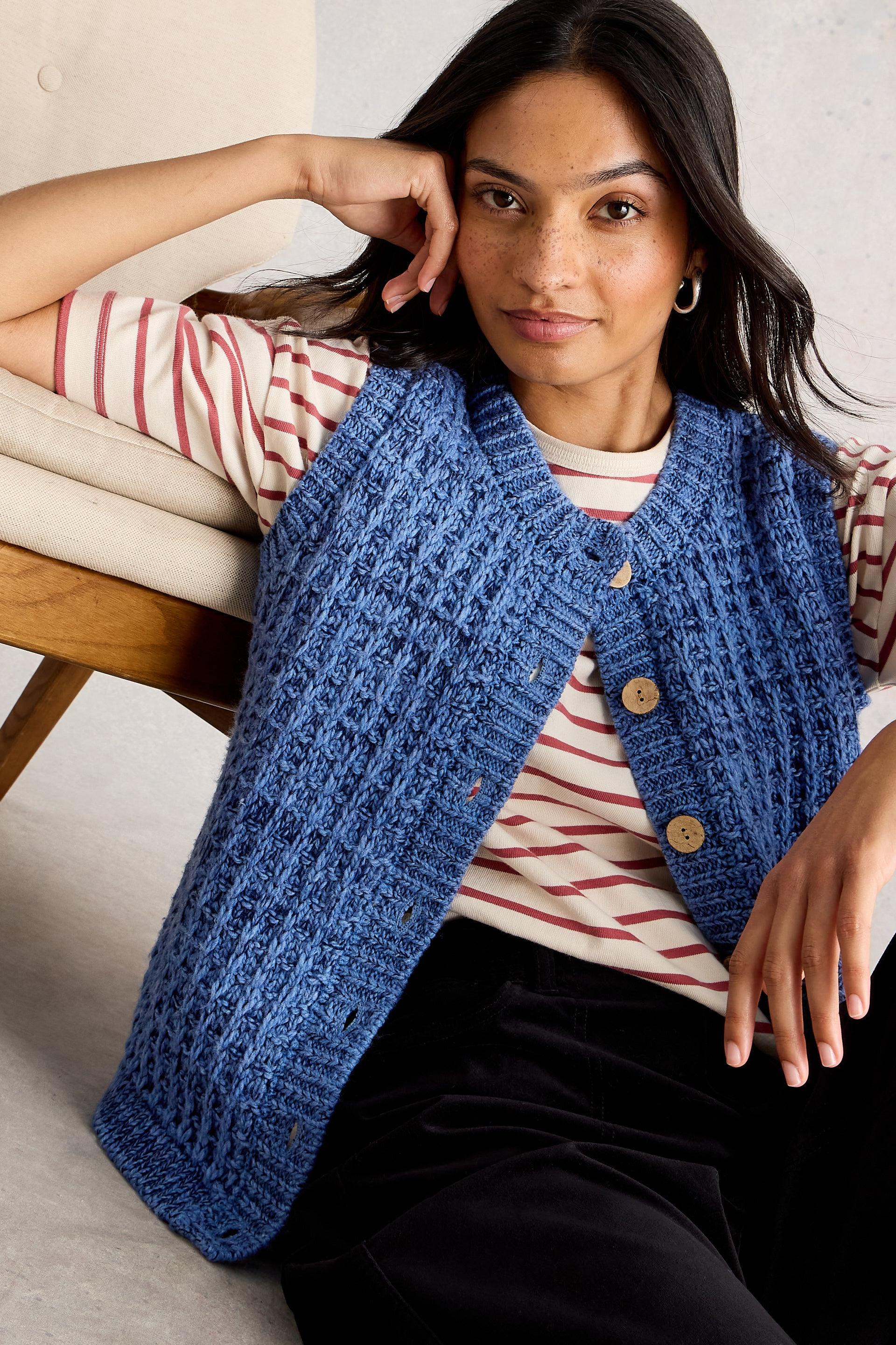 Woman wearing a blue knitted vest sitting against a chair.