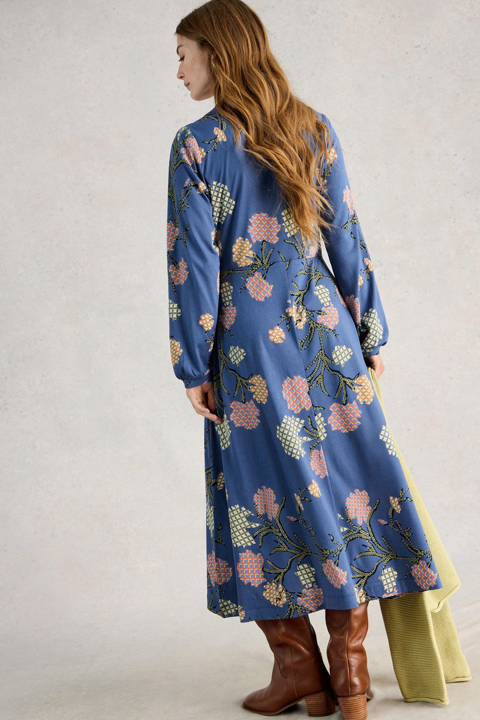 Woman wearing a blue floral organic cotton dress with brown boots on a plain background