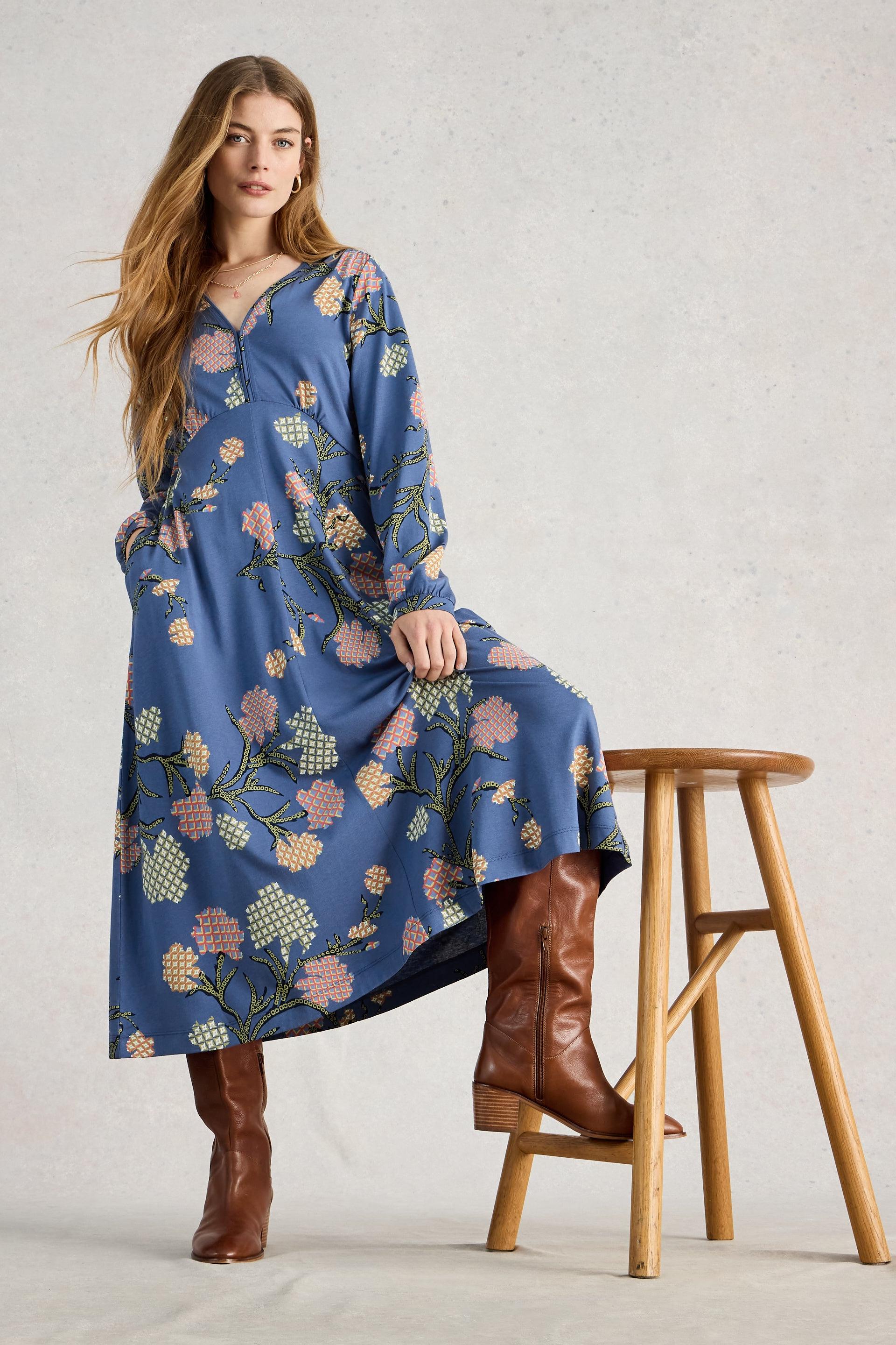 Woman wearing a blue floral dress and brown boots standing on a wooden stool against a light background