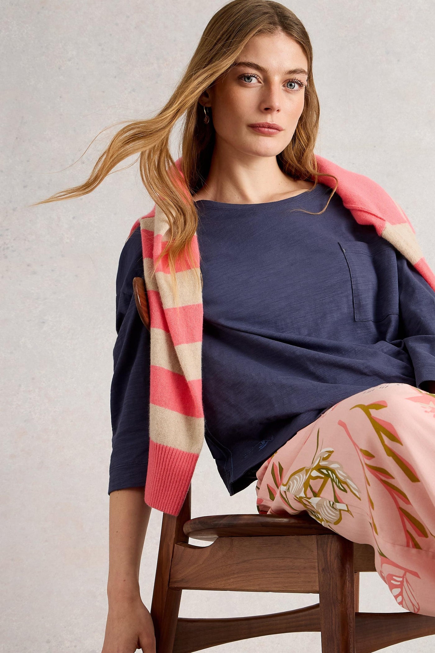 Woman sitting on a chair wearing a purple top and pink striped cardigan with floral pants.