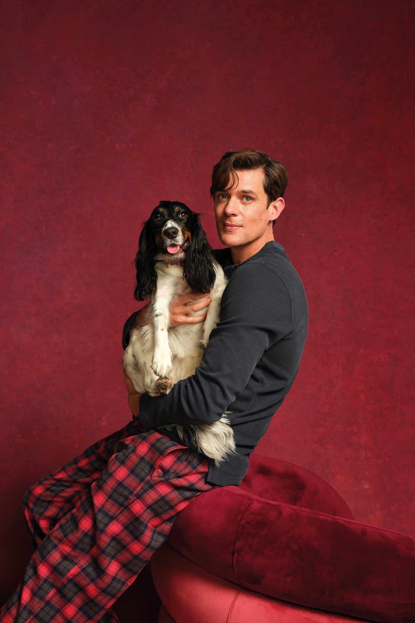 Man sitting on a red couch in organic cotton pyjamas holding a dog against a red background