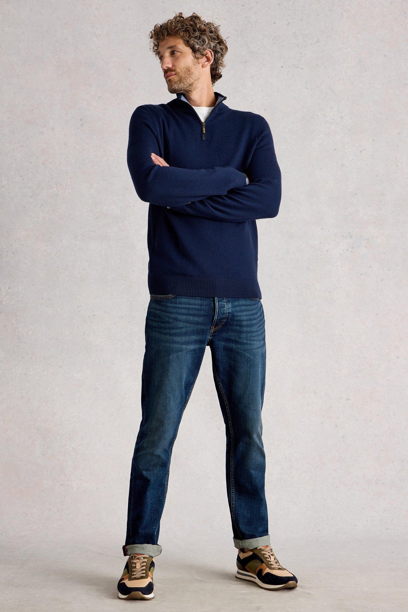 Man standing with folded arms wearing merino navy knit styled with blue denim jeans and cream and orange sneakers