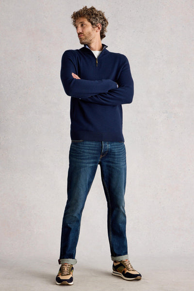 Man standing with folded arms wearing merino navy knit styled with blue denim jeans and cream and orange sneakers