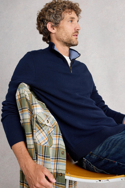 Man sitting on chair wearing navy merino wool long sleeve knit with quarter zip