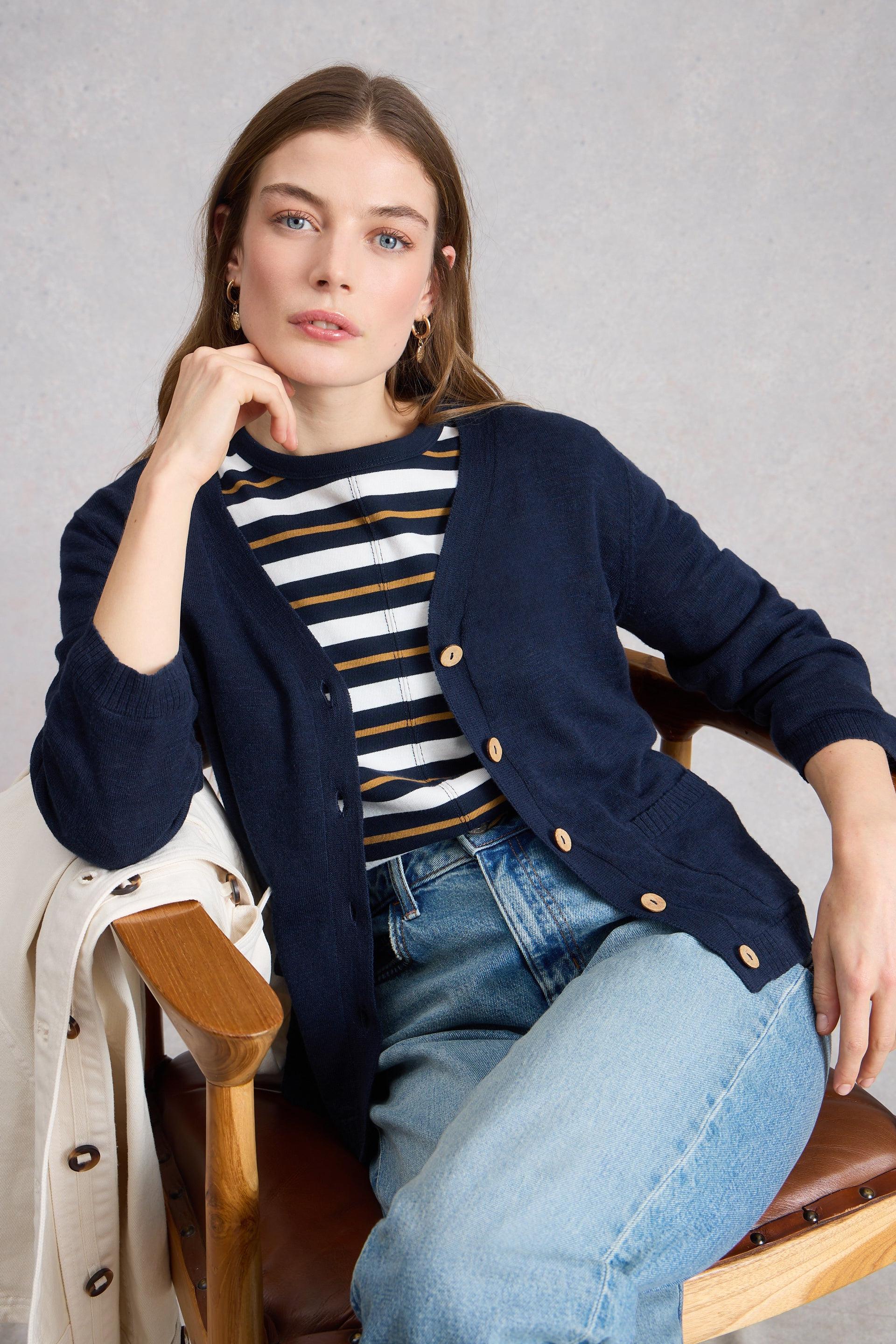 Woman wearing a navy organic cotton cardigan over a striped shirt and jeans, sitting on a chair against a gray background.