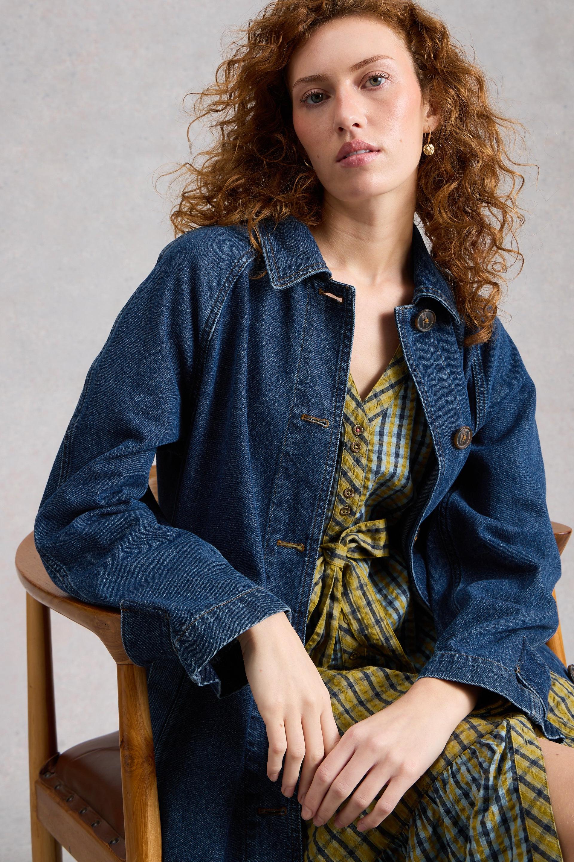 Woman wearing an organic cotton denim jacket over a patterned dress, sitting on a wooden chair against a plain background.