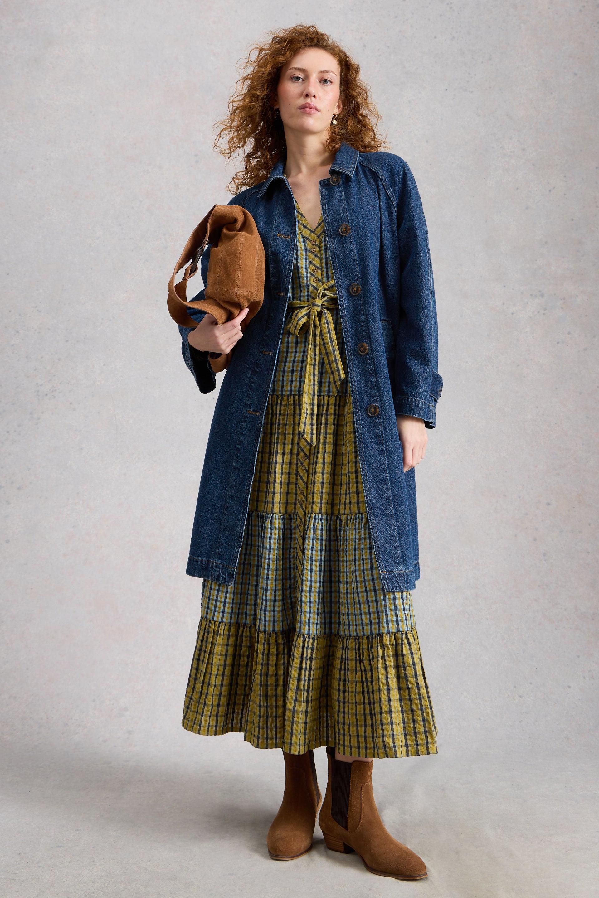 Woman wearing a blue organic cotton coat over a plaid dress holding a brown jacket against a plain background